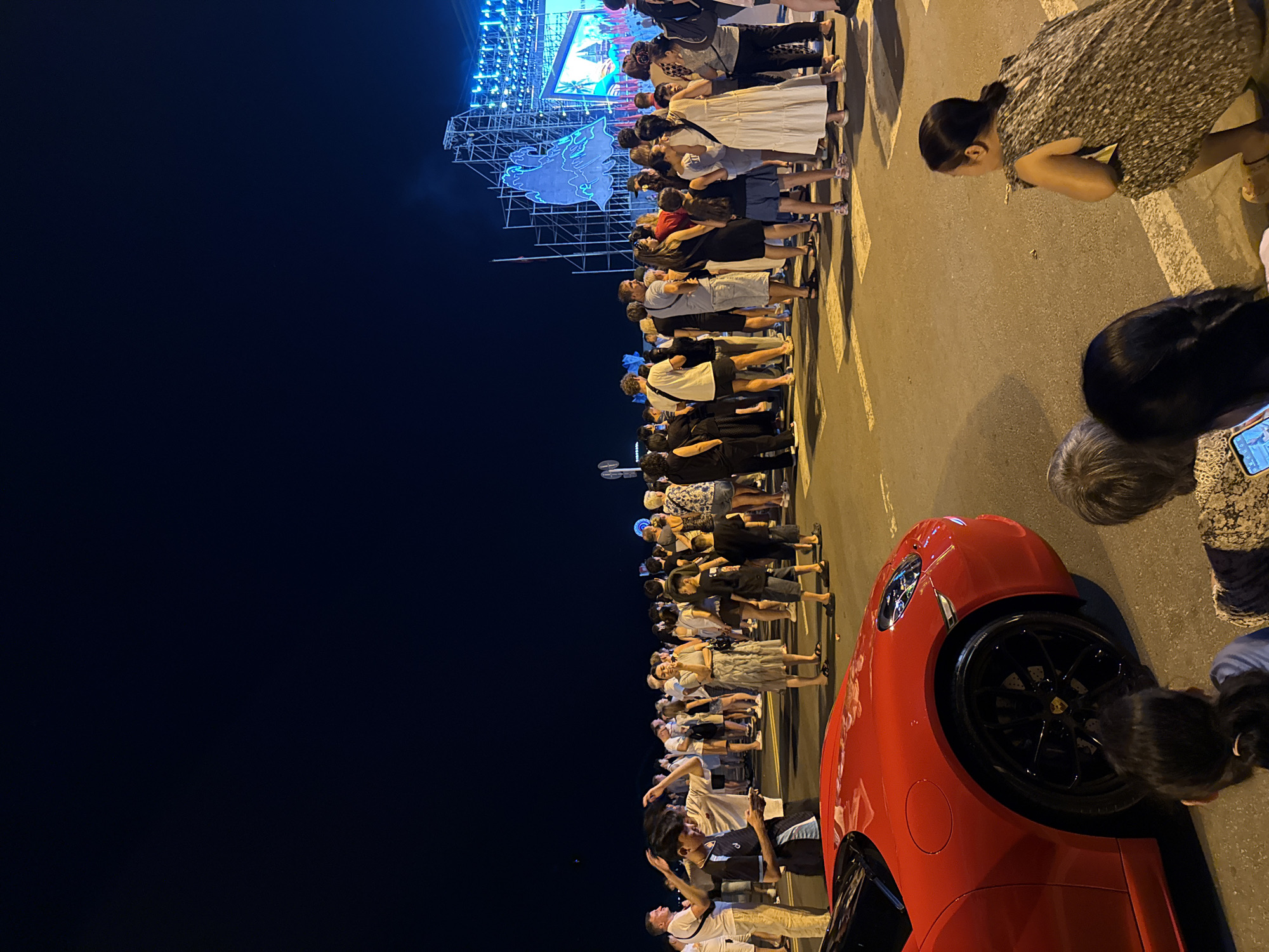 A nighttime scene depicts a crowded street in Nha Trang, Vietnam, possibly near Tran Phu Beach, with a bright, illuminated stage structure visible in the distance. A red sports car is parked on the left side of the frame, partially obscuring the foreground. A diverse crowd of people, including men, women, and children, are gathered, some standing, some sitting, and some appearing to be walking. The atmosphere suggests a public event or festival, with the stage lights illuminating the area. The street appears to be paved, with white pedestrian crossing lines visible. The sky is dark, indicating it is nighttime.
