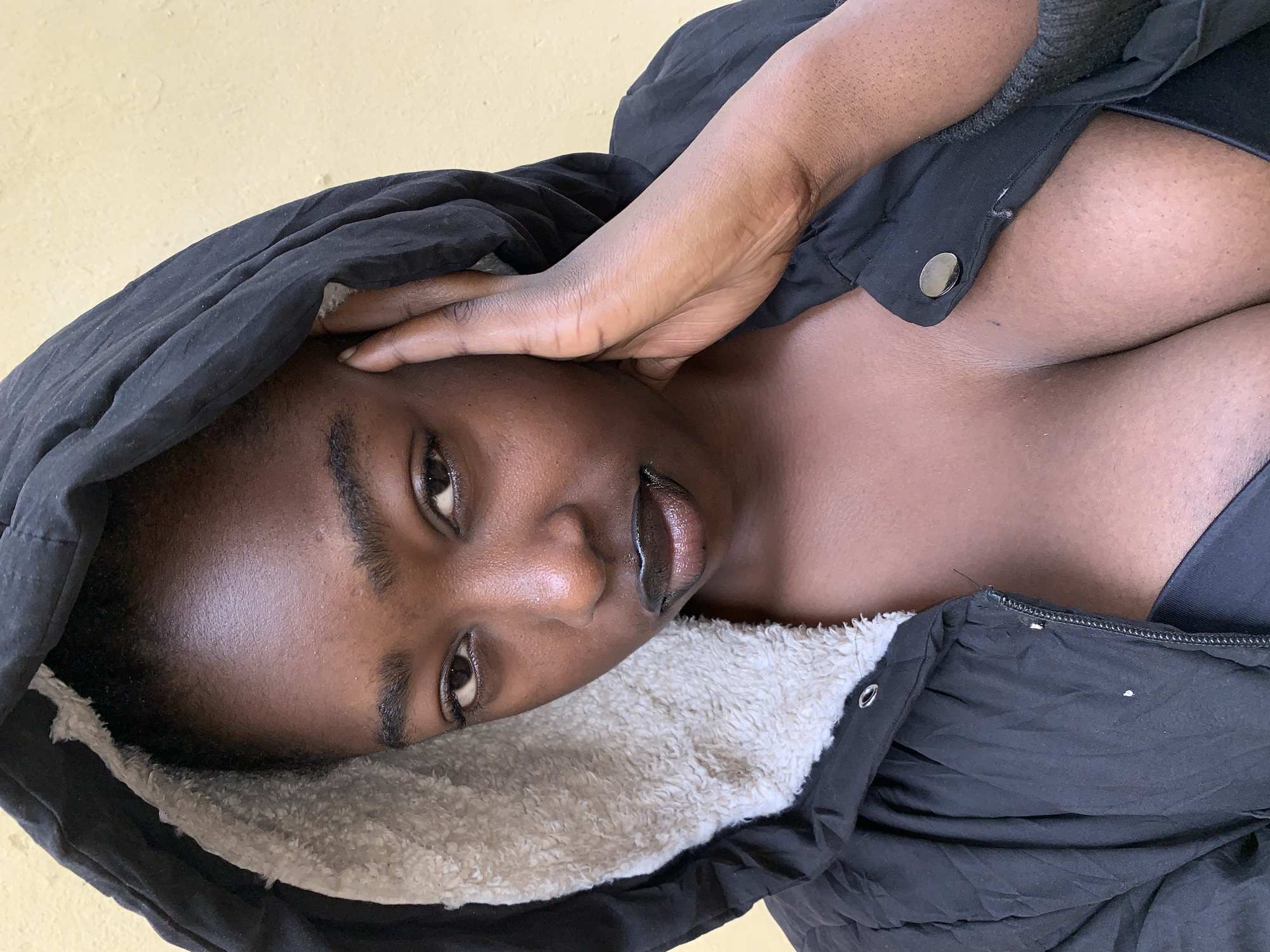 A close-up, angled shot shows a Black woman lying down, possibly indoors against a light yellowish wall. She is wearing a black hooded jacket with a white fleece lining. Her hand is touching her forehead, partially obscuring her face. The jacket is partially unbuttoned, revealing part of her upper chest and the straps of what appears to be an undergarment. It is not possible to confirm a specific location within Rafin Par, Nigeria from this image.