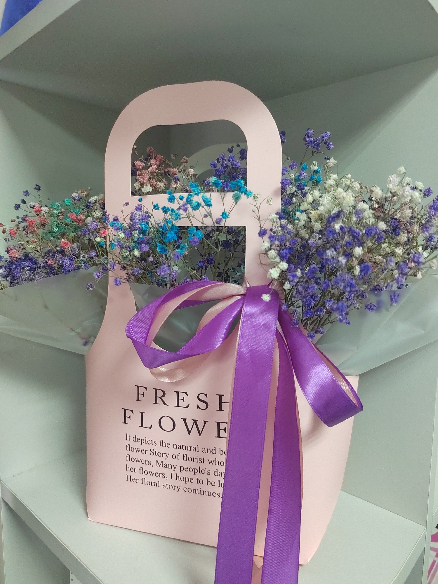 A pink floral gift bag sits on a light blue shelf, filled with multicolored baby's breath flowers. The bag has a handle at the top and is decorated with a purple ribbon tied in a bow. Text on the bag reads: "FRESH FLOWER" and then in smaller print, "It depicts the natural and b flower Story of florist who flowers, Many people's day her flowers, I hope to be h Her floral story continues.". The background is a light blue, slightly out of focus. The lighting is soft and appears to be artificial. The image is taken at an eye-level perspective. The scene is indoors, likely in a retail or storage environment. There is no indication of the specific location within the city of Biysk, Russia.