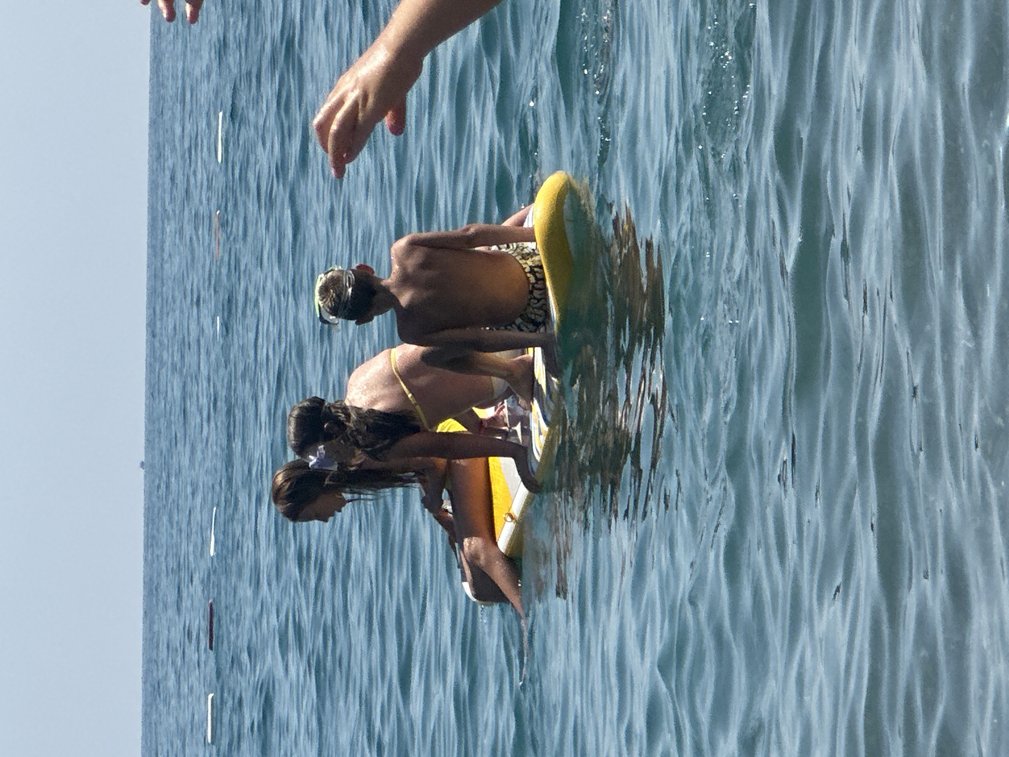 Four individuals are on a paddleboard in the sea. The scene is set in Boreti, Montenegro. Two young girls, with long brown hair, sit in the front. One girl is wearing a yellow bikini top and white bottoms. The other girl has her hair up with a purple accessory. Behind them, a young boy with a snorkeling mask on his forehead is seated. He is wearing patterned swim trunks. The paddleboard has a yellow and white striped pattern. The water is blue and slightly choppy, with a clear sky above. In the background, several white and red buoys are visible on the water. A distant boat can be seen on the horizon. The right side of the image shows the hands of two people reaching out.