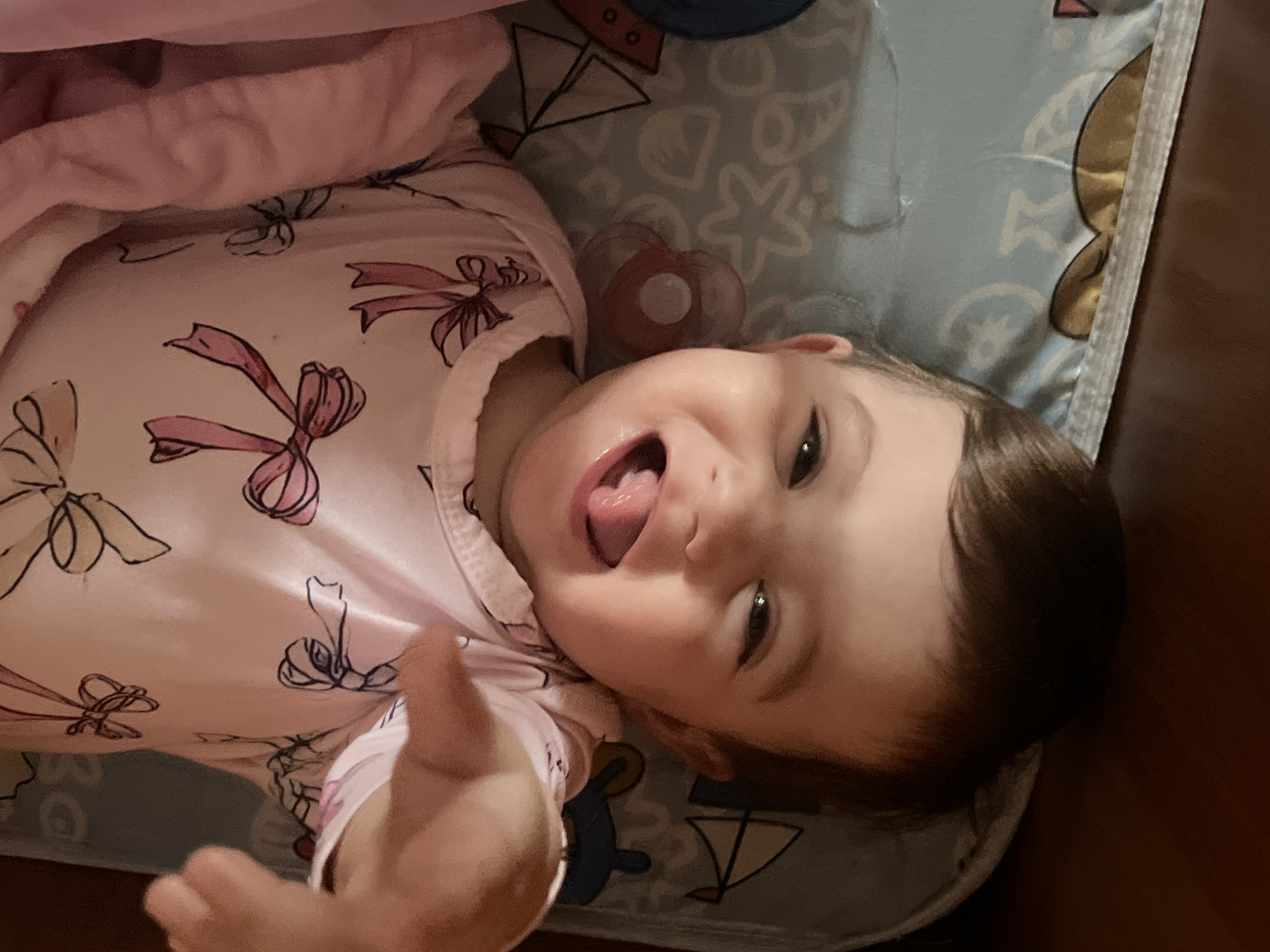 A baby lies on a patterned blue and white blanket, wearing a pink outfit with bow designs. The baby is looking at the camera, sticking its tongue out, and extending a hand towards the viewer. A pacifier lies near the baby's face. The scene is indoors, and the edge of a brown wooden surface is visible to the right. Elena, Bulgaria is not depicted in the image.