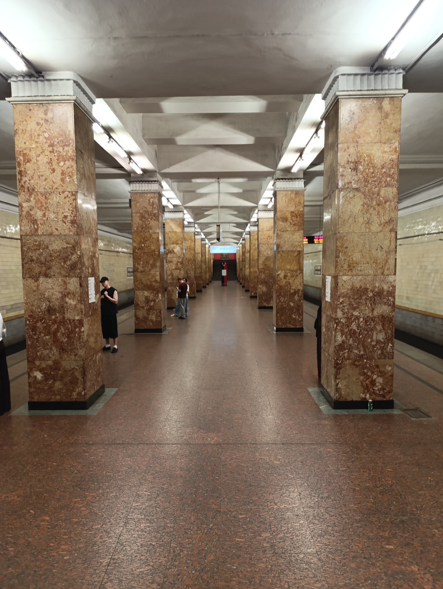 The image depicts the interior of a Moscow Metro station platform or passage, characterized by a long, symmetrical perspective. Numerous square columns, clad in reddish-brown variegated marble, line both sides of the passage, extending into the distance. Each column is topped with a white, fluted capital. The ceiling is high, off-white, with visible concrete beams and rows of fluorescent tube lighting providing illumination. The floor is composed of reddish-brown speckled terrazzo or tile, showing faint grout lines.
Along the left side, partially cropped, is a person in a white shirt. Further down, a male figure wearing a black sleeveless shirt, black shorts, and a black baseball cap stands facing a column, appearing to read a white paper or sign affixed to it. On the right side of the passage, in the mid-ground, a person in dark clothing is partially visible behind a column. Further down on the right, two individuals are present: one leans against a column wearing a light blue top and light trousers, while another, largely obscured, stands nearby. In the far distance on the right wall, a red digital display shows Cyrillic text and numbers, including "ИС", "02:10", and "22:32", likely indicating train information. A red cylindrical object, possibly a fire extinguisher stand or emergency beacon, is visible in the center of the passage further down. Near the base of the rearmost column on the right, a single green aluminum can rests on the floor. The walls behind the columns appear to be tiled in a light, possibly yellowish-beige hue.