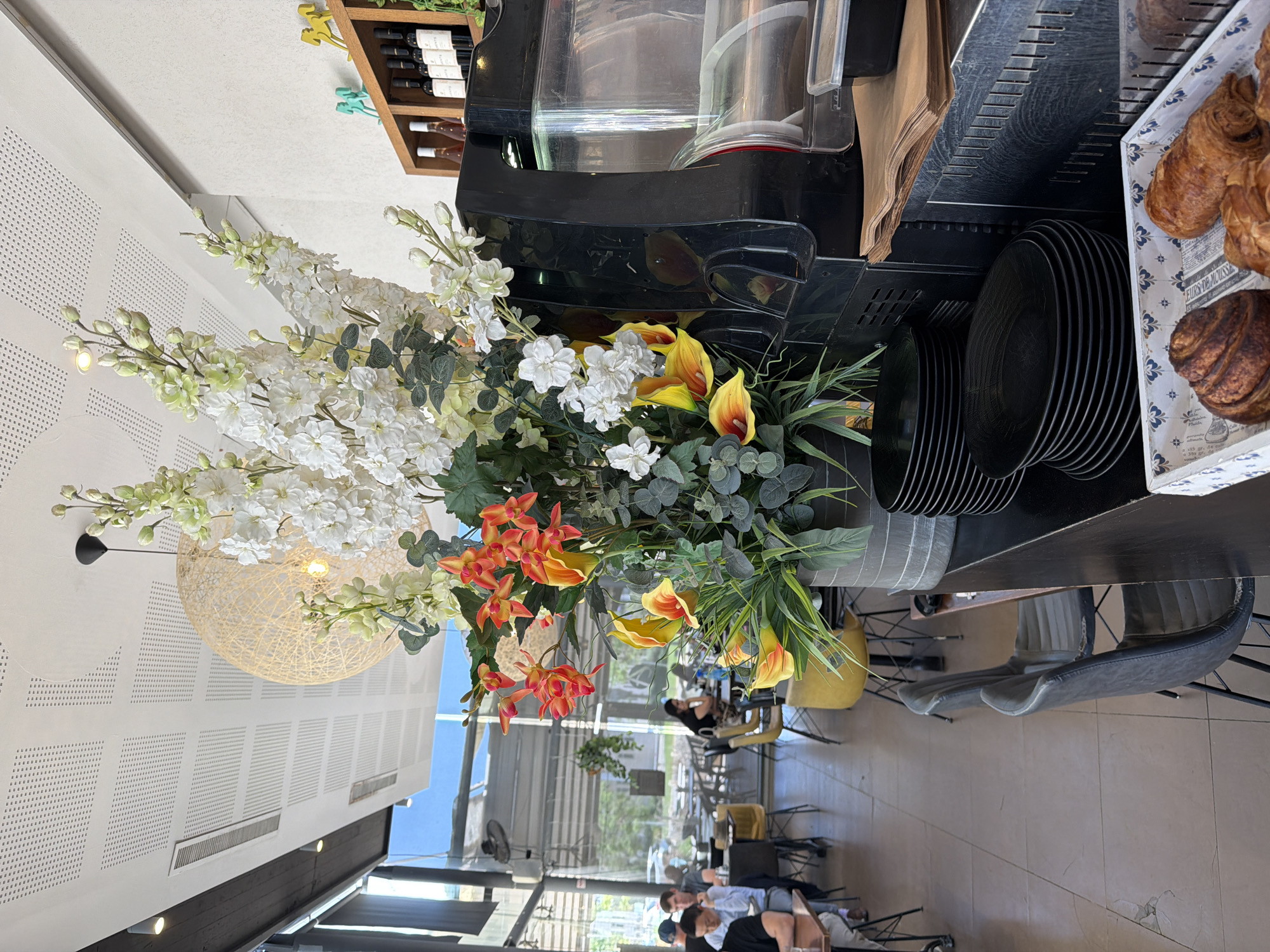 This image shows a still life arrangement of flowers and cafe items, with a blurred background of a cafe interior. In the foreground, a large bouquet of artificial flowers in white, orange, and yellow is prominently displayed. The white flowers are delicate and clustered, while the orange and yellow ones are more dramatic, resembling lilies or calla lilies. The flowers are arranged in a tall, dark vase or container.
To the right of the flowers, a stack of black plates is visible. Above the plates, a white box with blue patterns contains two croissants. There is also a glimpse of a coffee machine with a clear carafe and a black base. A brown paper napkin dispenser is also visible.
The background shows a cafe setting with tables and chairs. Several people are seated at tables, but their features are indistinct due to the blur. The overall lighting suggests it is daytime. The scene has a relaxed, casual atmosphere, typical of a cafe. There is no discernible text in the image. The arrangement of flowers and food items suggests a decorative display in a food service establishment. The image is taken from a slightly elevated angle, looking down and across the scene.