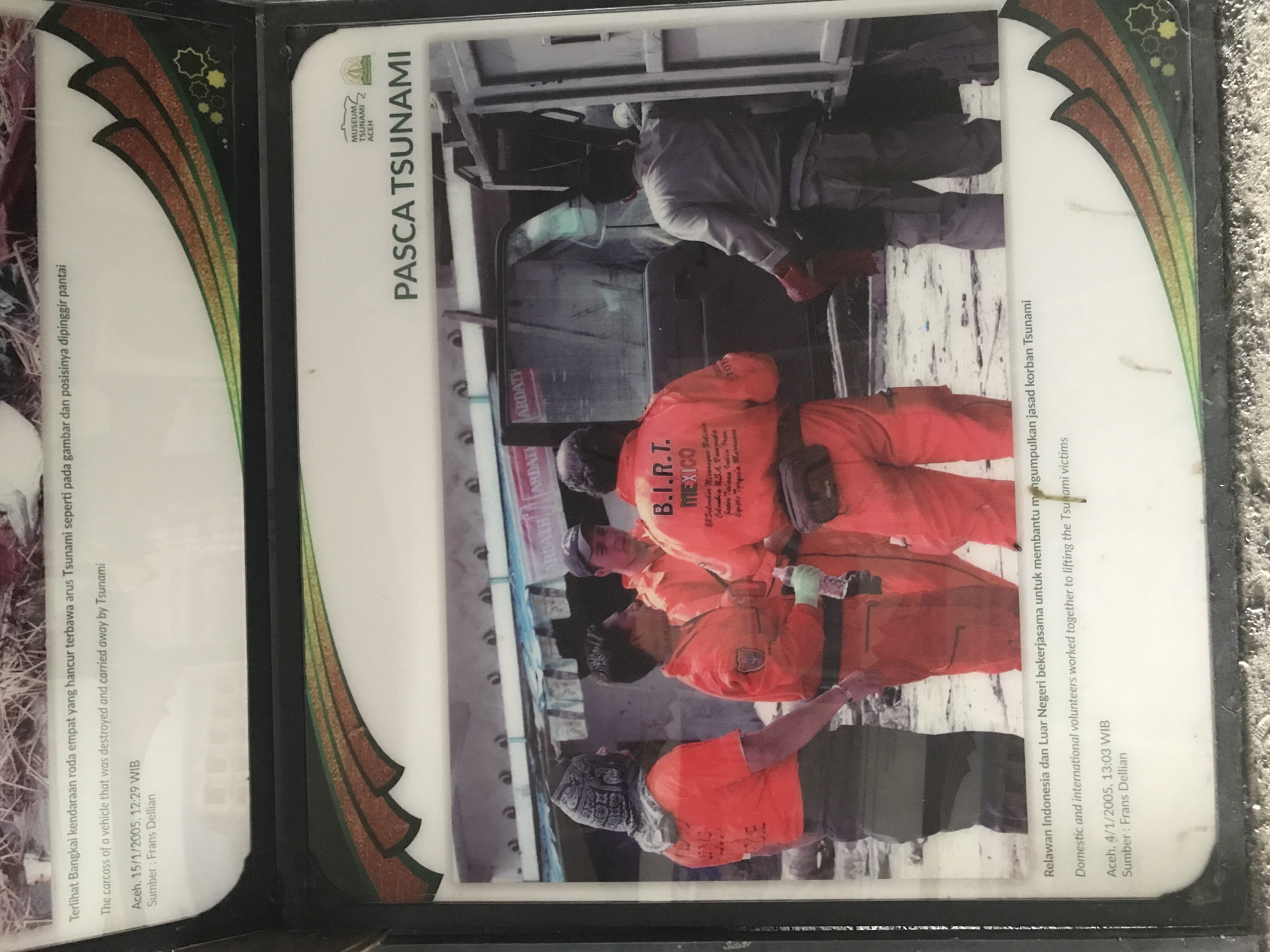 The image is a photograph of a museum exhibit depicting the aftermath of a tsunami. The title of the exhibit is "PASCA TSUNAMI," which translates to "Post-Tsunami." The photograph itself shows a group of people in orange jumpsuits identified as "B.I.R.T. Mexico" (likely a disaster response team). They appear to be working around a damaged vehicle, possibly retrieving bodies. The exhibit states that the people in the photograph are both domestic and international volunteers working together to lift Tsunami victims. A caption states that the carcass of a vehicle was destroyed and carried away by the Tsunami. The photograph of the vehicle carcass was taken on January 15, 2005, at 12:29 WIB in Aceh, Indonesia. The photograph of the disaster relief team was taken on January 4, 2005, at 13:03 WIB, also in Aceh, Indonesia.