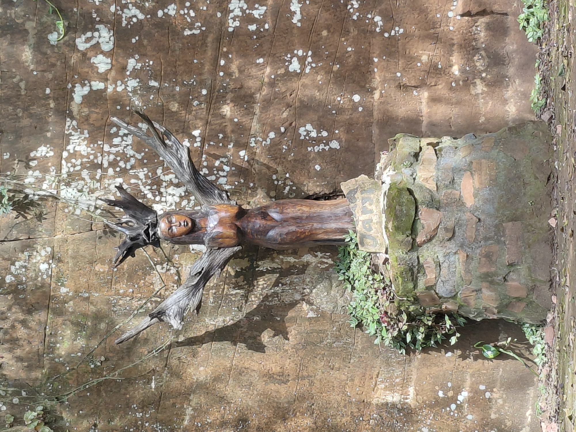 A wooden statue of a female figure with outstretched arms is displayed in front of a textured stone wall. The statue is carved from dark wood, depicting a woman with detailed facial features and a slender body, wearing what appears to be a headdress made of driftwood. The statue stands on a pedestal constructed from rough stones and bricks, surrounded by greenery. The stone wall behind the statue is rough and weathered, with natural striations and lighter colored mineral deposits. The lighting suggests it is daytime, with shadows cast on the wall. The overall scene evokes a sense of natural artistry and integration with the environment. The location is identified as being in the city of Trinidad, Paraguay.
