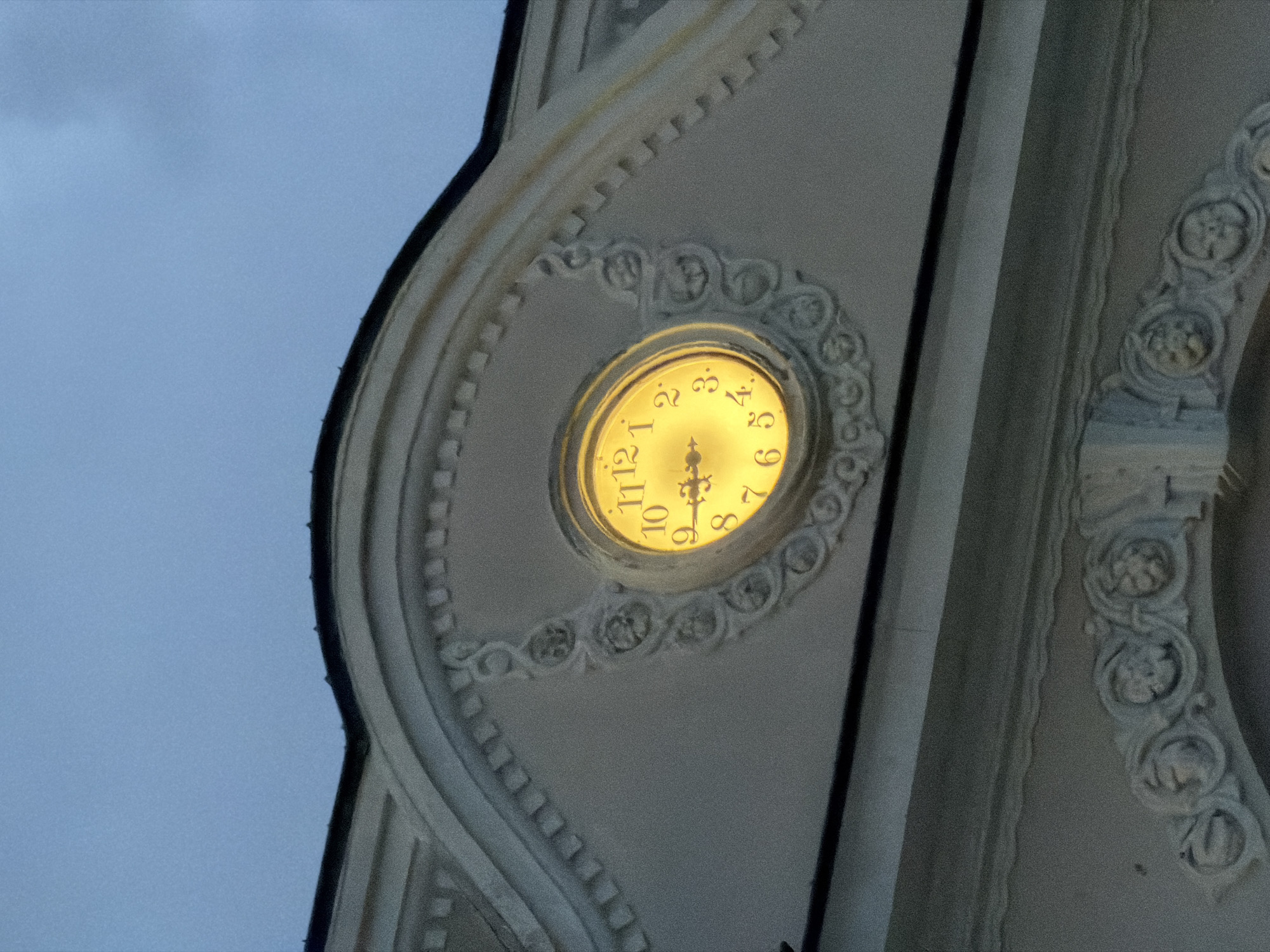An upward-angled, close-up shot captures a section of an ornate building in Blagoevgrad, Bulgaria, highlighting an illuminated clock face. The clock, glowing with a warm yellow light, is prominently centered in the frame. The time displayed on the clock is approximately 8:58 or 9:00, with the minute hand pointing towards the twelve and the hour hand positioned just before the nine. All twelve numbers on the clock face are visible in a stylized serif font.
The clock is embedded within rich architectural detailing, including intricate white or light-grey carvings, moldings, and a partial view of a column or pilaster to the right. The decorative elements feature elaborate scrollwork, floral patterns, and what appear to be small sculptural elements or busts, indicative of classical or baroque design. The building material has a textured appearance, likely stone or stucco.
The background shows a soft, muted blue-grey sky, suggesting it is either twilight or nighttime, which explains the internal illumination of the clock face. The image emphasizes the detailed craftsmanship of the historical or significant building against the evening sky.