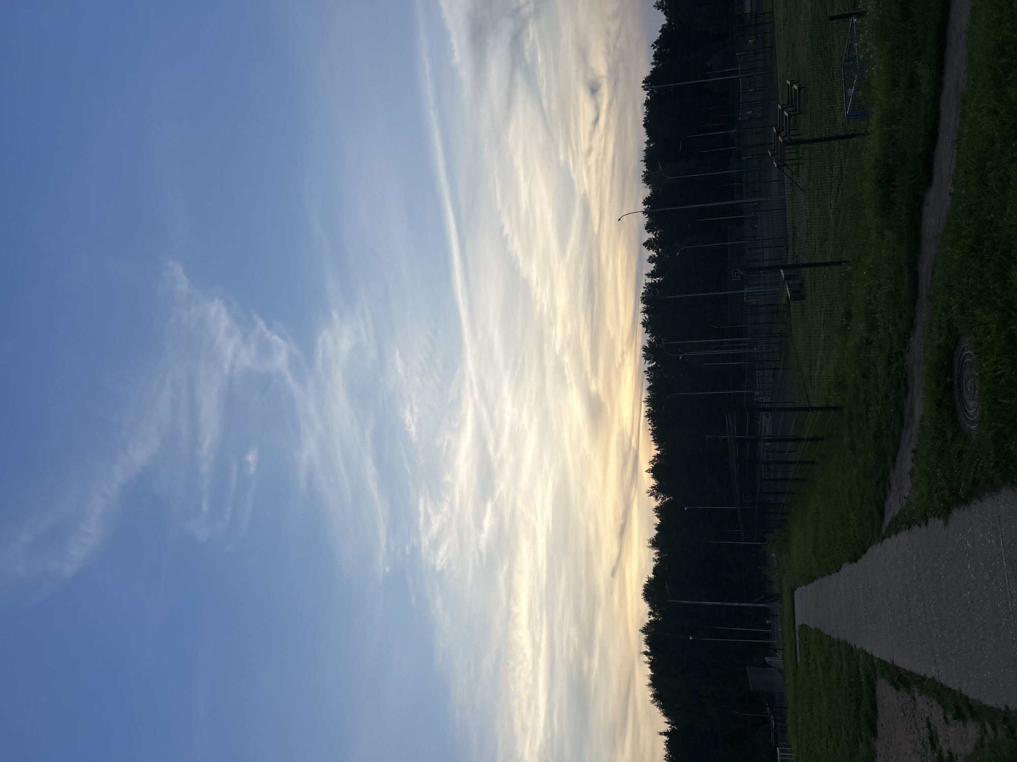 The image depicts an outdoor scene at dusk, likely in the city of Марковский, Russia. In the foreground, a paved pathway leads through a grassy area. To the right of the path, a fenced sports field is visible, containing what appears to be playground equipment. Beyond the field, a dense line of dark trees forms a treeline. Above the trees, the sky is a gradient of colors, transitioning from a pale yellow and orange near the horizon to a clear blue at the top, with wispy clouds scattered across it. Streetlights are present along the edge of the treeline, some of them illuminated. The overall impression is a peaceful, possibly recreational area at the end of the day. There are no people visible in the image.