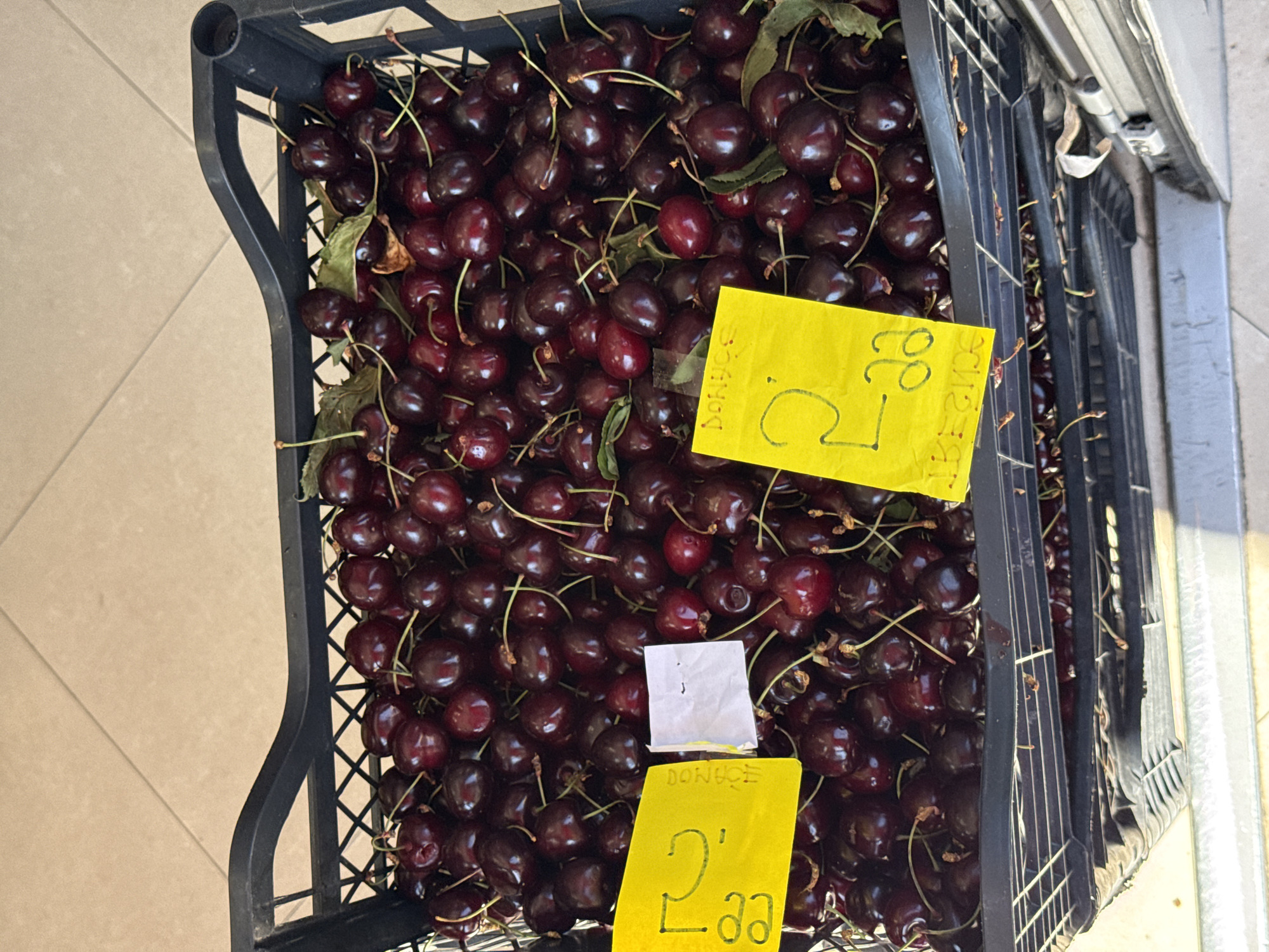 A close-up, high-angle shot shows a black plastic crate overflowing with ripe, dark red cherries. Two yellow price tags are visible in the crate. One tag, on the left, displays "2, 99". The other tag, on the right, also shows "2, 99" and has the word "DONUGE" written above it. A small white piece of paper is also tucked among the cherries on the left. The crate is placed on a tiled surface, with the floor tiles visible at the top of the image. The bottom of the image shows the ribbed texture of the crate and a glimpse of a metal railing. The scene appears to be an outdoor market stall, possibly in Budva, Montenegro.