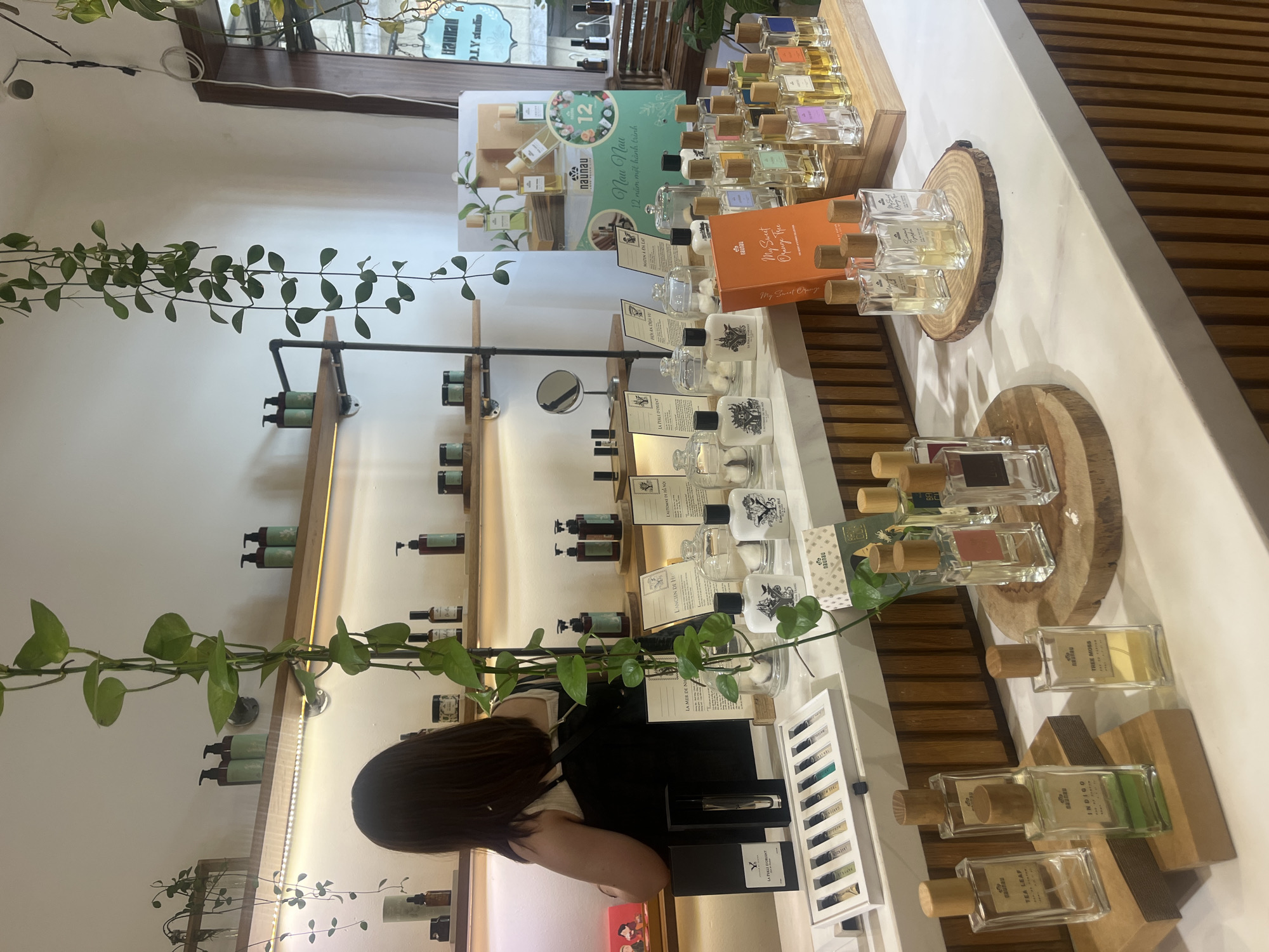The image depicts the interior of a retail store specializing in perfumes and scents, likely located in Ho Chi Minh City, Vietnam. The store features a minimalist aesthetic with clean white walls and wooden shelving. Hanging plants add a touch of natural greenery to the space.
In the foreground, a white counter displays a variety of perfume bottles. Several glass bottles with wooden stoppers are arranged on small wooden platforms. Some bottles have labels such as "TEA LEAF," "INDIGO," and "TREE MOSS," indicating different fragrances. To the left, a set of small perfume sample vials is laid out in a rectangular tray. In the center, an orange box with the text "Nau Nau My Sweet Orange Tree" is visible. Behind these, a larger selection of perfume bottles of various sizes and colors are arranged on tiered wooden shelves.
A woman with dark hair is partially visible in the background, standing behind the counter and facing away from the camera, appearing to be attending to customers or organizing products. She is wearing a white top and a dark apron or bag.
The shelves in the background are stocked with more products, including bottles with pumps, jars, and decorative items. A large promotional poster for "Nau Nau" with images of perfume bottles and text in Vietnamese is displayed on the right side of the back wall. The overall impression is of a well-organized and aesthetically pleasing retail environment.