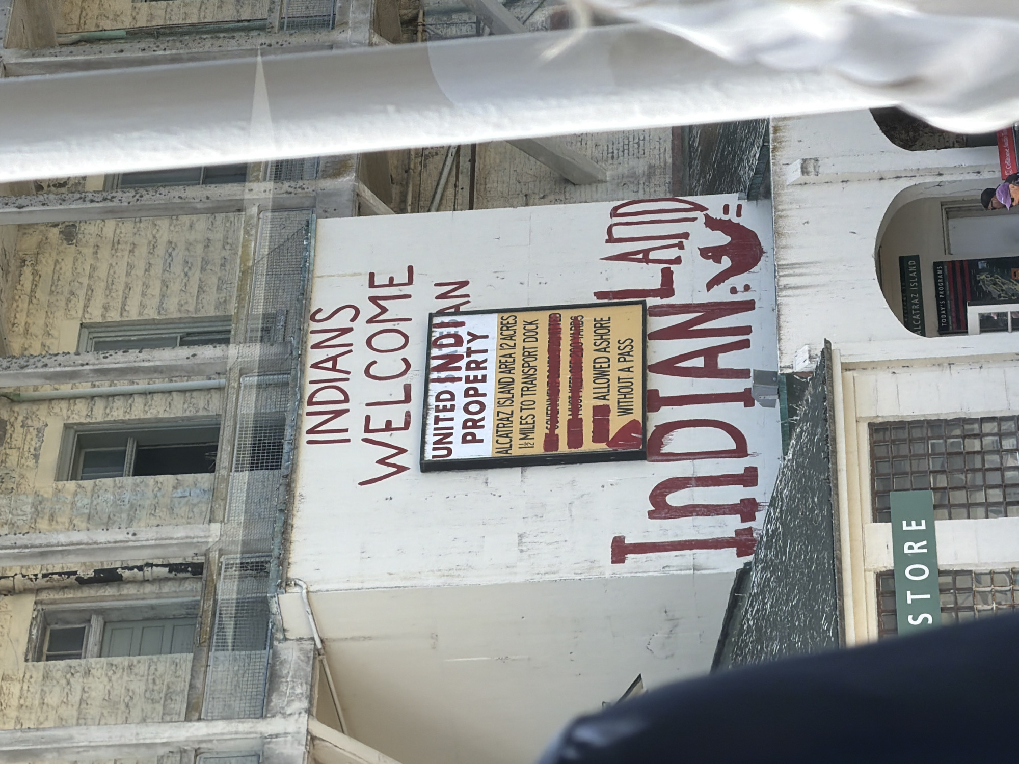 The image depicts a sign reading "INDIANS WELCOME" in large red letters on the side of a white building. Below this, a smaller sign is visible with text such as "UNITED INDIAN PROPERTY" and information about Alcatraz Island, including "ALCATRAZ ISLAND AREA 12 ACRES" and "1/2 MILES TO TRANSPORT DOCK." Further down the wall, a large red painting of a bird and the words "INDIAN LAND" are visible. In the foreground, a green sign that says "STORE" is mounted above a building with large, barred windows. The overall scene appears to be Alcatraz Island, a former federal prison located in San Francisco Bay, California.
