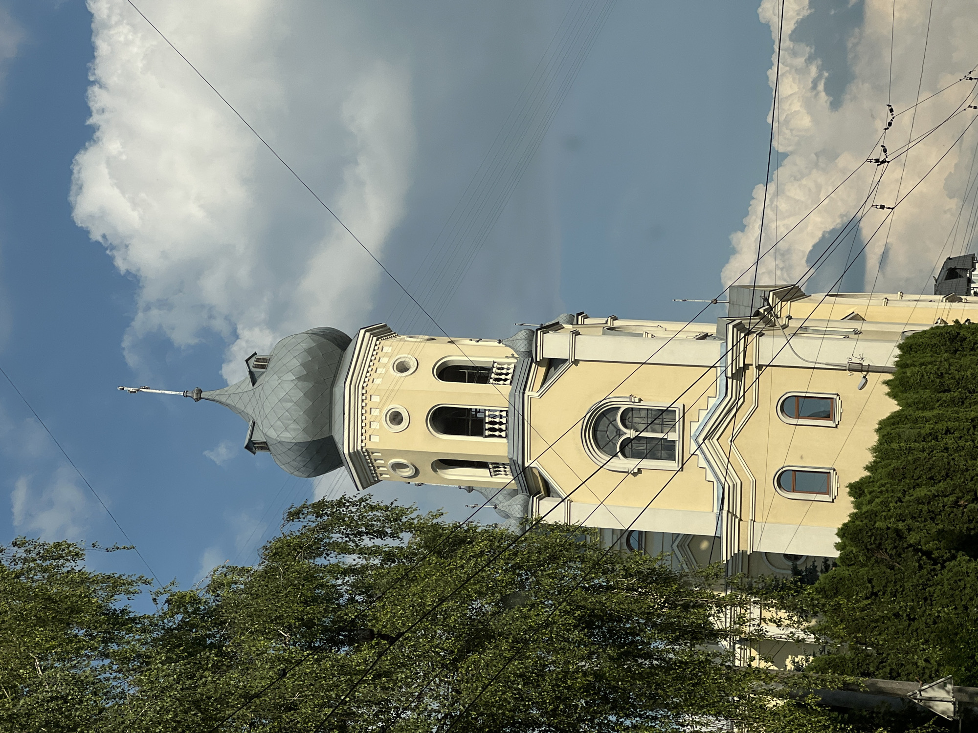 The image captures a close-up view of a yellow church tower with a grey onion dome, set against a backdrop of a blue sky with scattered white clouds. The tower features arched windows, circular openings, and decorative moldings. A large green tree with delicate leaves partially obscures the left side of the tower. Power lines crisscross the sky in front of the building. In the foreground, a dense line of dark green evergreen trees forms a hedge. The overall scene suggests an urban environment, possibly in Ternopil, Ukraine, given the architectural style common in Eastern Europe. There are no people or animals visible in the image.