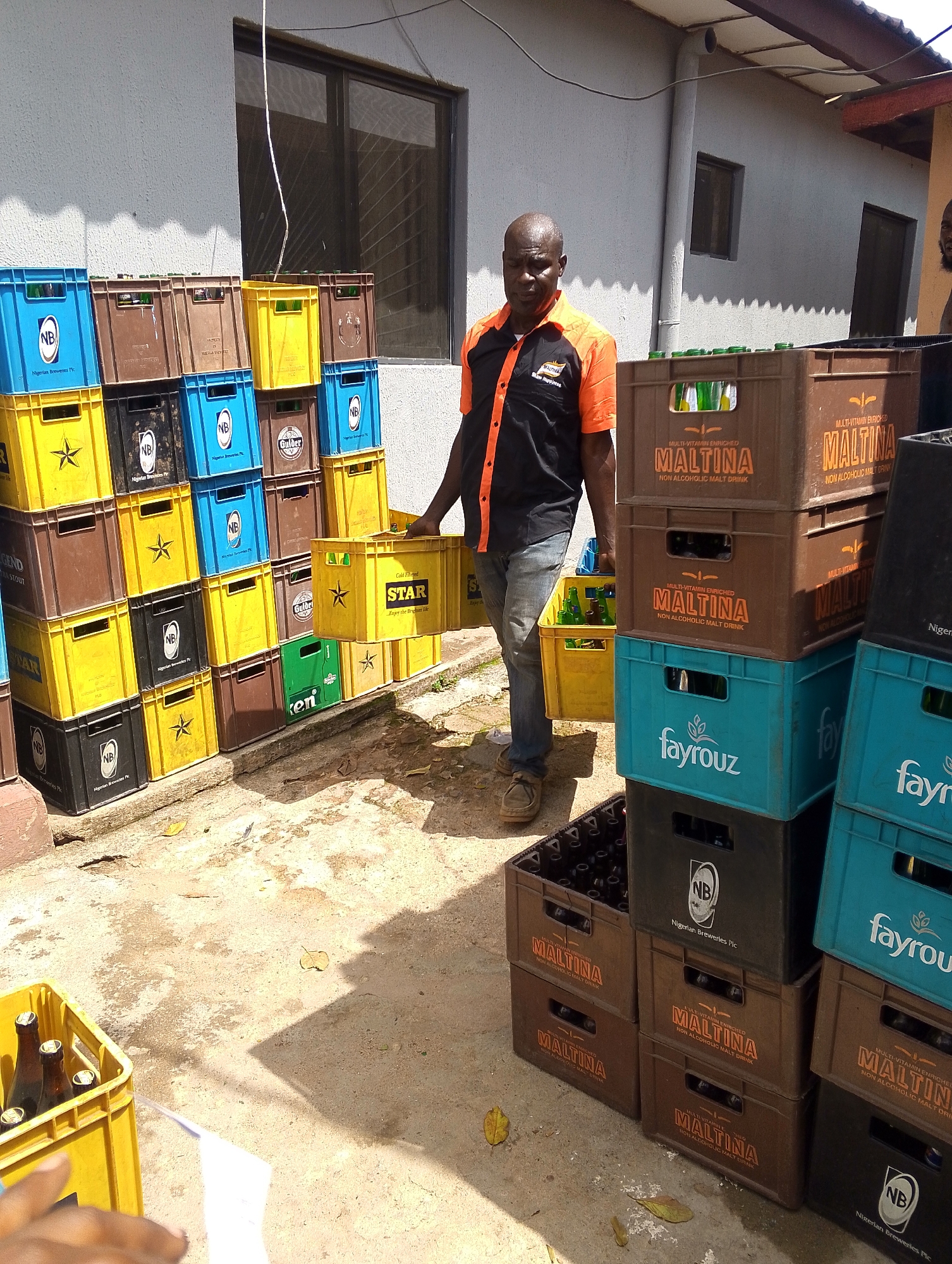 The image shows an outdoor scene in Jos, Nigeria, featuring a man carrying a crate of beer. The man, wearing a black and orange shirt and jeans, is walking towards the right side of the frame. Stacks of crates of various beverages, including "Maltina," "Fayrouz," "Star," "Gulder," and crates with "NB" markings (Nigerian Breweries), are arranged against a light gray building. Some crates are full of bottles. The ground is concrete with scattered leaves and debris. A partial view of another person's face is visible on the right side of the frame, along with a hand holding a white piece of paper at the bottom left corner. The building has windows with screens.