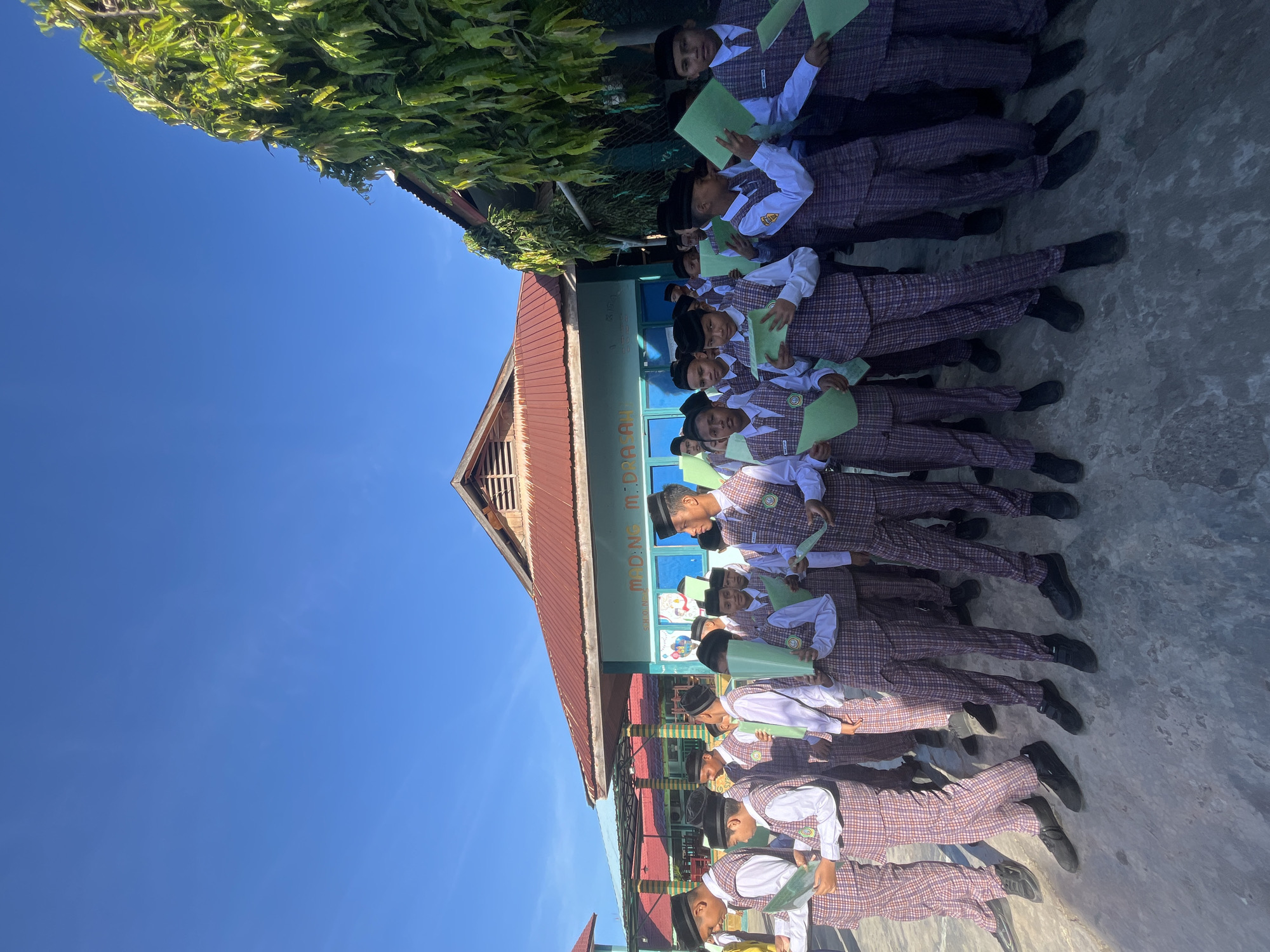 A group of young boys, dressed in plaid vests, white shirts, plaid pants, and black skullcaps, stand in a line outdoors, holding green papers. The boys appear to be students at a school, as suggested by the building in the background. The building has a red roof and light blue walls with signage. A large tree with green foliage is visible to the right of the building. The scene is illuminated by bright sunlight under a clear blue sky. The ground is paved with concrete. The location is identified as a school in Lhokseumawe, Indonesia.
