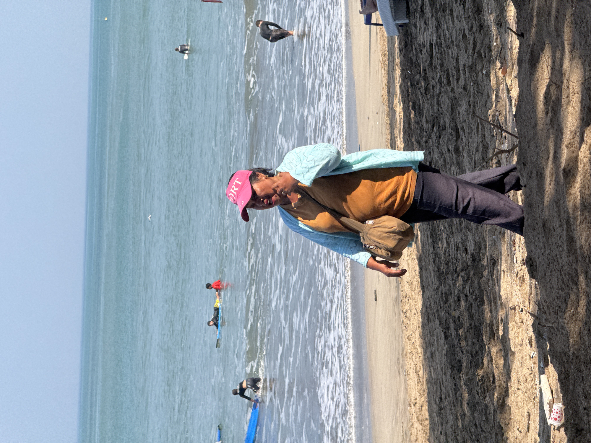 In Kuta, Indonesia, on a sunny day, a woman with a pink visor walks on a sandy beach near the ocean. She wears a brown shirt, a light blue cardigan, and dark pants. A light brown purse is slung over her shoulder. In the background, several people are in the water, some on surfboards. The ocean is calm with gentle waves breaking on the shore. The beach is covered in sand with some scattered debris and shadows from unseen trees.