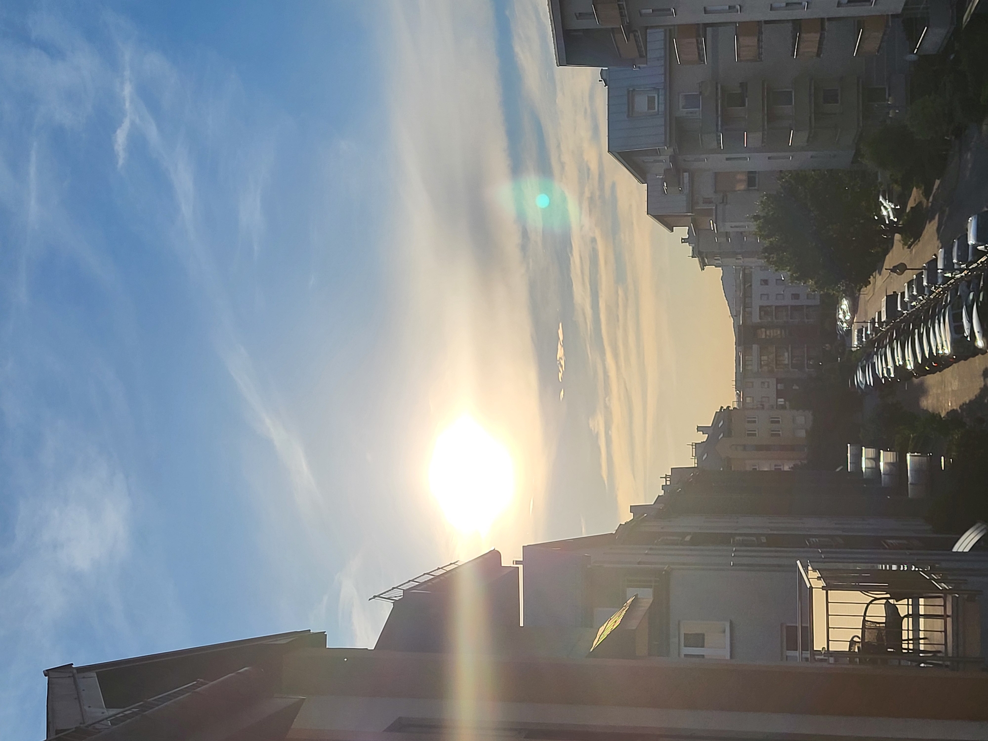 The image depicts a cityscape during what appears to be sunset or sunrise. The sun is a prominent bright orb in the sky, casting a warm glow and creating lens flare effects. The sky is a vibrant blue, with thin, wispy clouds that are illuminated by the sun.
Dominating the scene are multi-story buildings, likely apartment complexes, lining a street. The architecture suggests an urban or suburban setting. The buildings have visible windows and balconies, some of which are adorned with railings or small awnings. On the street level, a row of parked cars is visible, mostly light-colored sedans, suggesting a residential area or a parking lot. Trees are interspersed among the buildings, adding a touch of greenery to the urban landscape.
The perspective of the image suggests it was taken from an elevated position, looking down and across the street. The lighting indicates a time of day when the sun is low on the horizon, creating long shadows and a warm, golden hue on the buildings and sky. The overall mood is tranquil and serene, capturing a quiet moment in the urban environment as day transitions to night or vice versa. There is no visible text in the image.