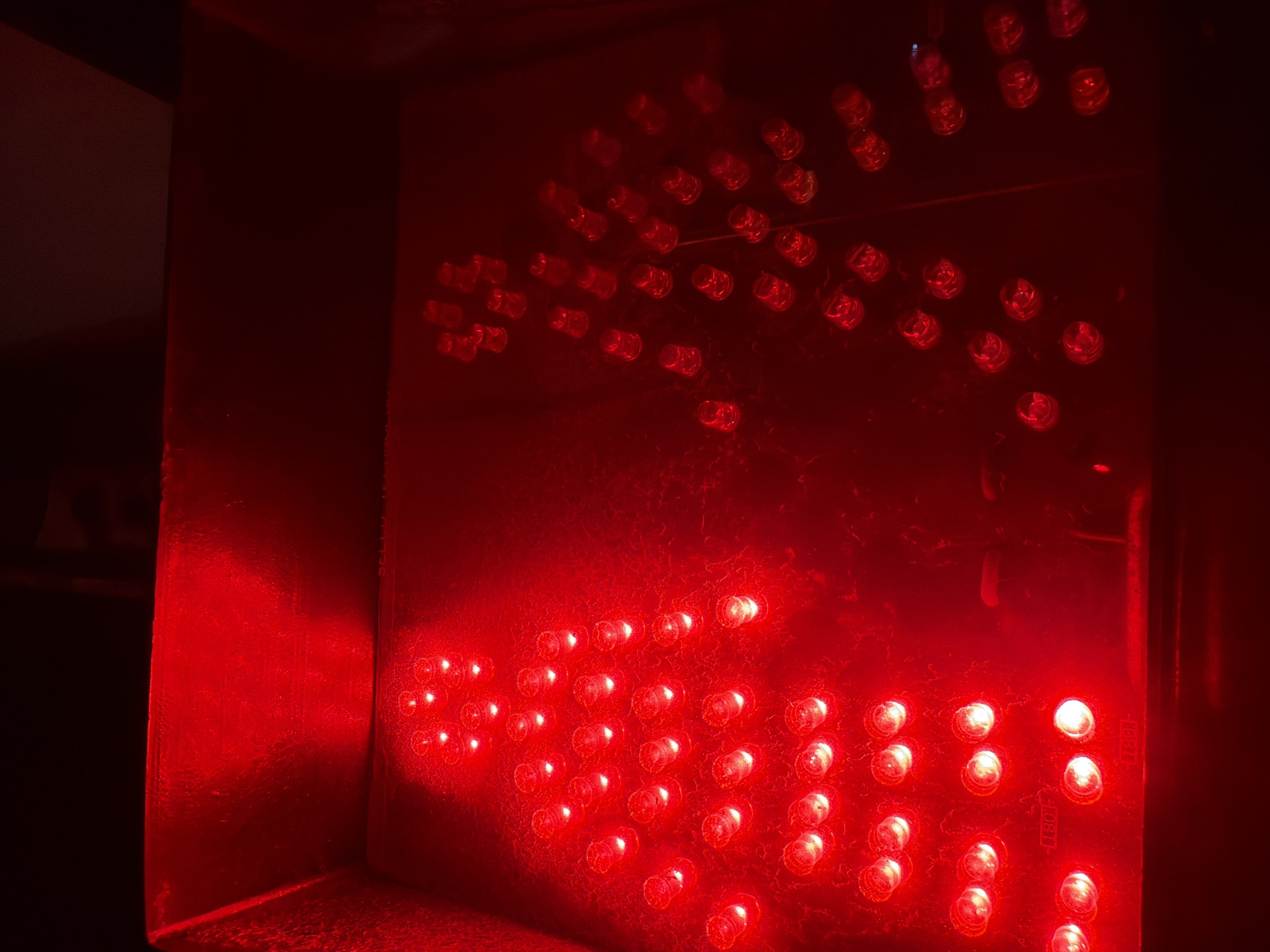 The image displays a close-up, overhead view of a red LED pedestrian crossing signal, illuminating a silhouetted human figure pointing to the right. The signal is within a dark, possibly indoor environment, with the red light casting reflections on the textured surface of the device and surrounding structure. Two distinct LED arrays form the figure: one on the left is more clearly defined and appears to be in focus, while the one on the right is slightly out of focus and partially obscured. The overall impression is of a functional traffic signal, possibly in a state of disrepair or being tested, given the isolated nature of the illuminated figure. The context provided of Nha Trang Ward, Vietnam, suggests this might be a street-level or building-integrated signal, though the image itself does not contain any explicit identifying landmarks of that location.