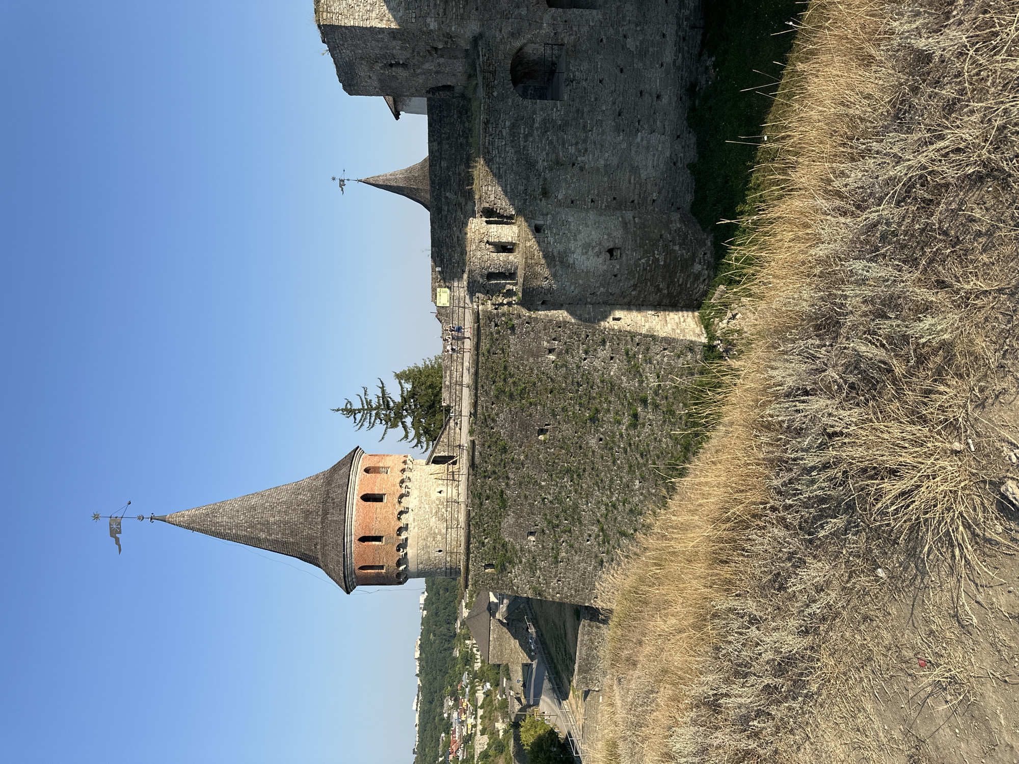 This image shows a view of the Kamianets-Podilskyi Castle in Kamianets-Podilskyi, Ukraine, on a clear, sunny day. The castle's imposing stone walls and a distinctive conical tower with a weather vane are prominent. The foreground is filled with dry, golden grass and sparse vegetation. In the mid-ground, the castle's stone ramparts and battlements are visible, with a few small trees growing on them. A walkway with a railing runs along the top of the ramparts, and several figures, likely tourists, are standing on it. In the background, a picturesque town with red-tiled roofs sprawls across a green, hilly landscape under a bright blue sky. The architecture suggests a historical fortress.