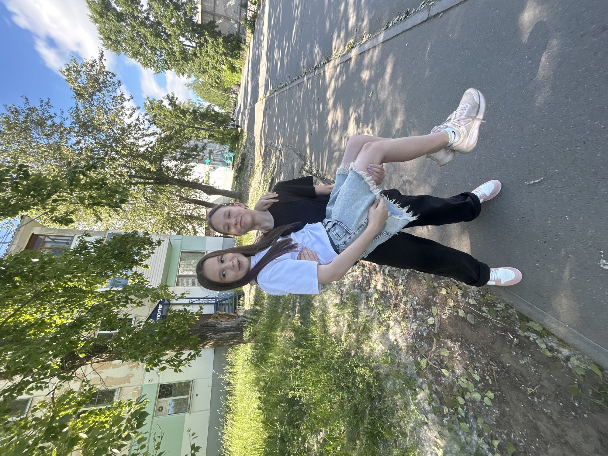 In Chaikovsky, Russia, on a sunny day, a young person carries another in their arms on a sidewalk next to a tree-lined street. The person carrying is wearing a black t-shirt and black pants, with pink and white sneakers. The person being carried is wearing a white t-shirt, a denim skirt, and pink sneakers. They are standing in front of a multi-story building with a green and white facade, and a tree with green leaves partially obscures the view. A paved sidewalk runs alongside the street, with grass and trees on the other side of the sidewalk. The street is asphalt and appears to be in good condition, with some parked cars visible in the distance. The sky is blue with scattered clouds.