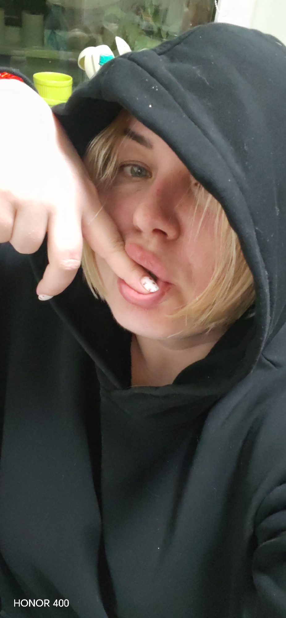 The image shows a person wearing a black hooded sweatshirt. The person's face is visible, with blond hair partially obscuring the forehead. One hand is raised, with a finger near the mouth. The fingernails are painted with a design. The background is blurry and includes hints of other objects. The text "HONOR 400" is visible in the bottom left corner. The location is not specified.