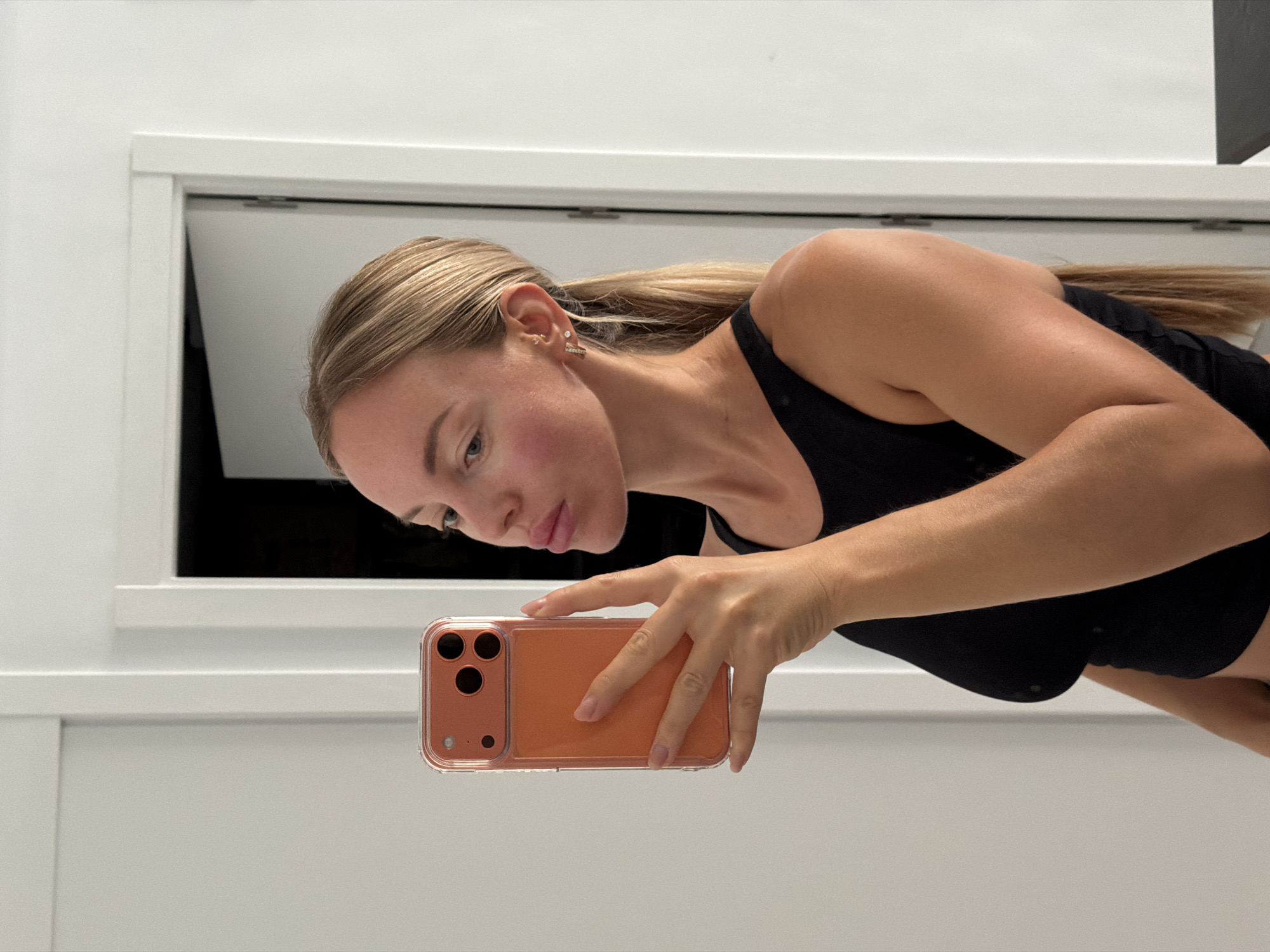 A medium-shot, eye-level, profile shot shows a fair-skinned woman with blonde hair pulled back in a ponytail. She is wearing a black tank top and is holding a cell phone in her right hand, taking a selfie in a mirror. The woman is in a room with white walls and a white door in the background. The scene is indoors. No information regarding the location in the city of Finestrat, Spain is available in the image.