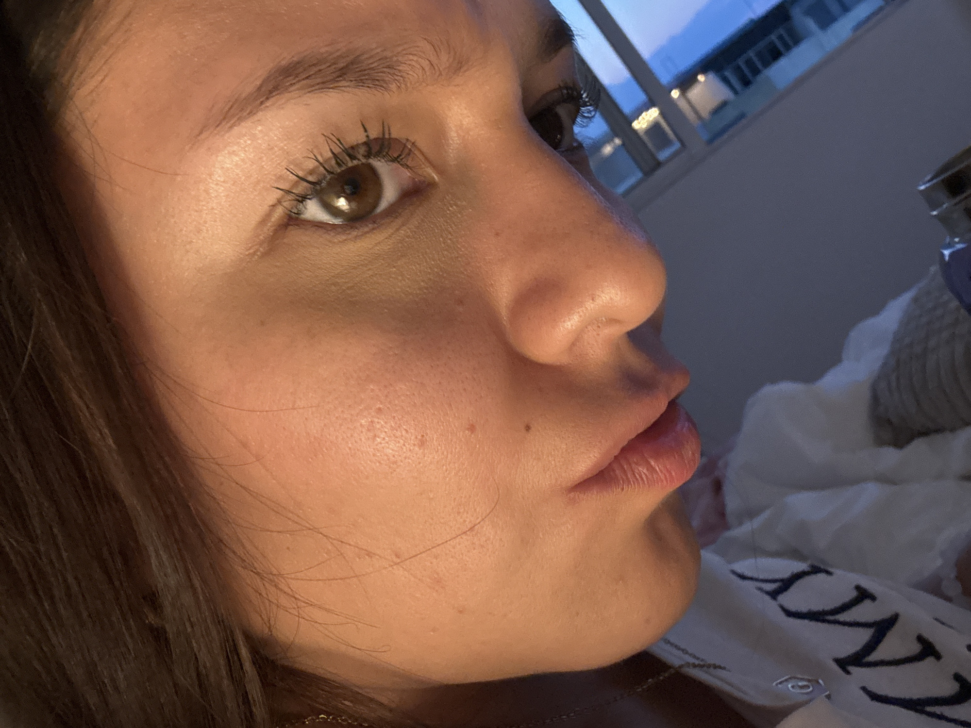 A close-up, angled portrait of a young woman with brown eyes, dark brown hair and light skin. She has mascara on her upper lashes. Her lips are slightly pursed. A small mole is visible on her left cheek. The background is blurred, showing a window with the sky visible and a white pillow with lettering. The location of the image is not discernible from the provided image. It is impossible to determine if the location is San Francisco Ocotlán (Ocotlán), Mexico based on the image details.