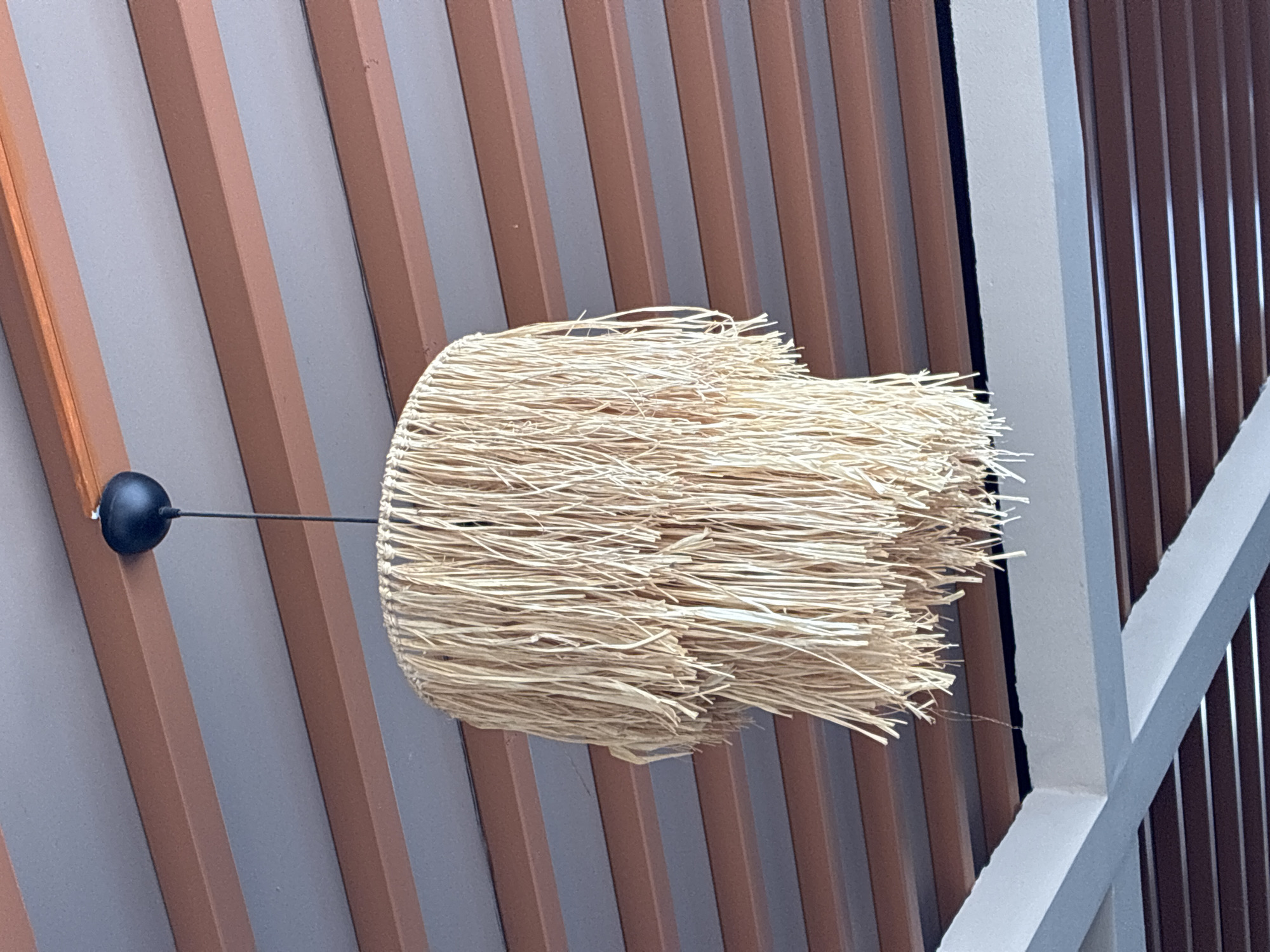 A bohemian-style hanging lamp made of natural raffia or straw dangles from a ceiling. The lamp has multiple tiers of fringe that create a textured and layered effect. Above the lamp, the ceiling is constructed with alternating wooden beams and grey panels. The scene appears to be an interior or covered outdoor space, possibly a restaurant, cafe, or covered patio area. The lighting suggests it could be daytime, with natural light filtering from an unseen source. This lamp could be found in a variety of settings in Alanya, Turkey, from trendy cafes in the city center to beachside restaurants along the Mediterranean coast.
