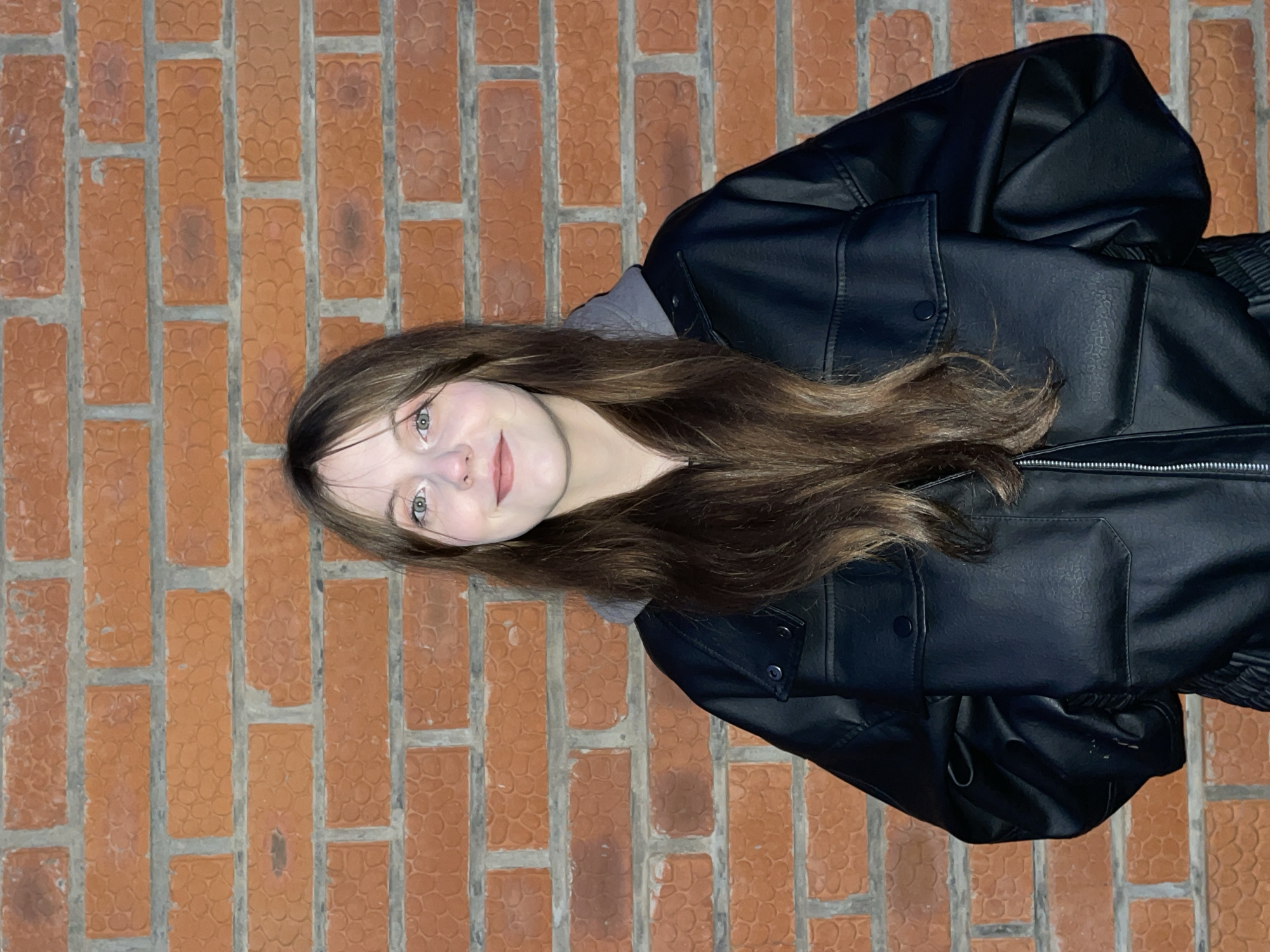 A young woman with long brown hair stands in front of a brick wall. She wears a dark leather jacket over a gray hoodie. The setting is unclear and cannot be specified as Фоки, Russia, based on the visual information provided.