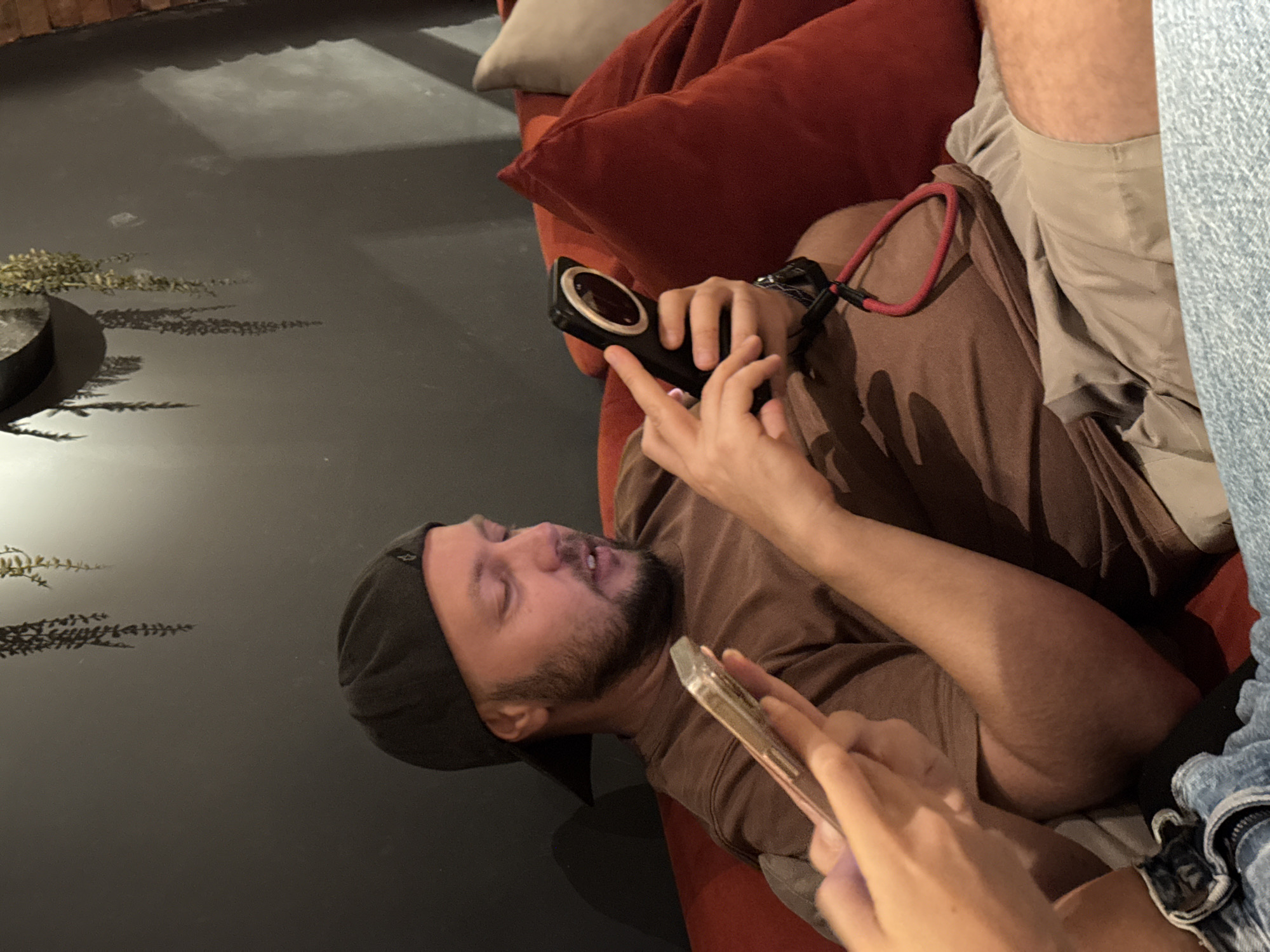 This is a media file of a person lying on a red couch, possibly in a lounge area. The person is wearing a brown t-shirt and a dark baseball cap, with a beard. They are holding a black smartphone with a circular lens. Another person's hands are visible in the foreground, holding a transparent phone case. The setting appears to be indoors with a dark floor and a hint of greenery in the background, possibly plants. The lighting suggests it might be evening or a dimly lit indoor space. The overall mood seems relaxed, with the person on the couch looking engaged with their phone. The context indicates this might be Hà Nội, Vietnam. There is no visible text in the image.