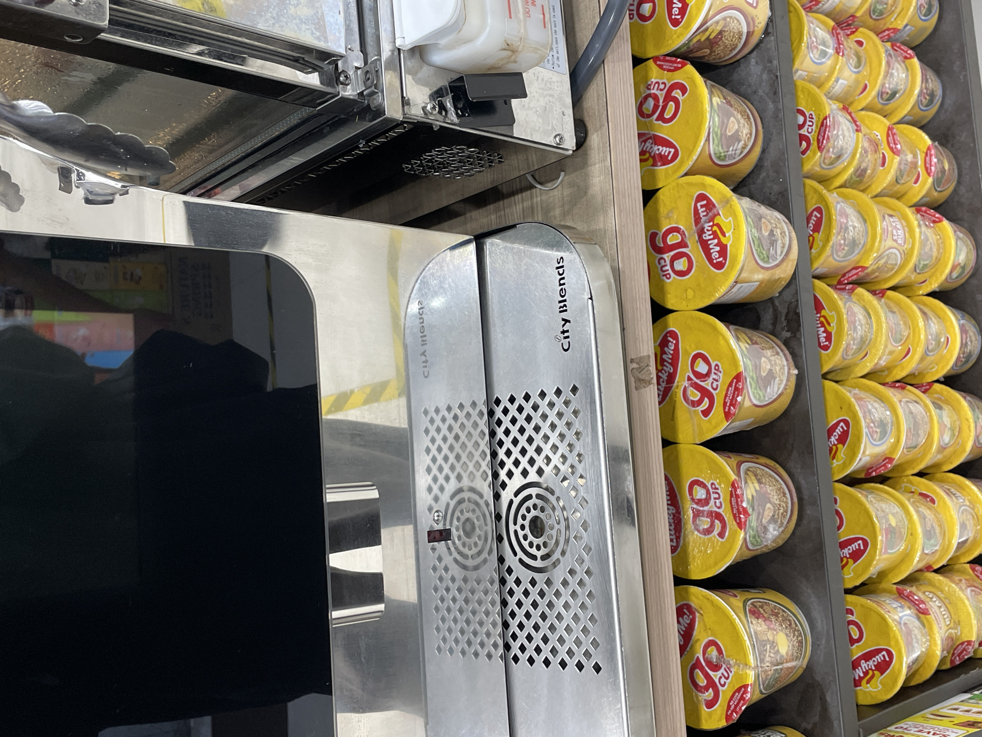 The image captures an indoor scene, likely a food service area, featuring a commercial stainless steel counter and shelves stocked with instant noodle cups. On the right side, multiple rows of yellow-colored instant noodle cups are neatly arranged. The most prominent brand visible is "Lucky Me! Go Cup," with some cups also showing "BULALO," indicating a flavor.
To the left, a gleaming stainless steel counter or appliance is visible, with a pair of metal tongs resting on its reflective surface. Adjacent to this counter, a silver metallic device is prominently displayed, labeled "City Blends." This device features a perforated, grid-like design with circular vents, and a small red indicator light. Mirrored text, "CILA BIGUCI2," is also visible on this device, which appears to be a reflection of "City Blends." Above the "City Blends" device, a smaller white container is attached to another piece of stainless steel equipment. Partially visible text on this container includes phrases like "DO NOT," "IN," "Follow," and "of the unit, once the unit is cool." A large black rectangular surface, possibly a screen or part of another appliance, occupies the lower left portion of the image.
The overall impression is that of a clean, well-organized quick-service food station, likely where customers can select instant noodles and potentially prepare them using hot water from a dispenser like the "City Blends" unit. The lighting is bright and even, consistent with an indoor commercial setting.