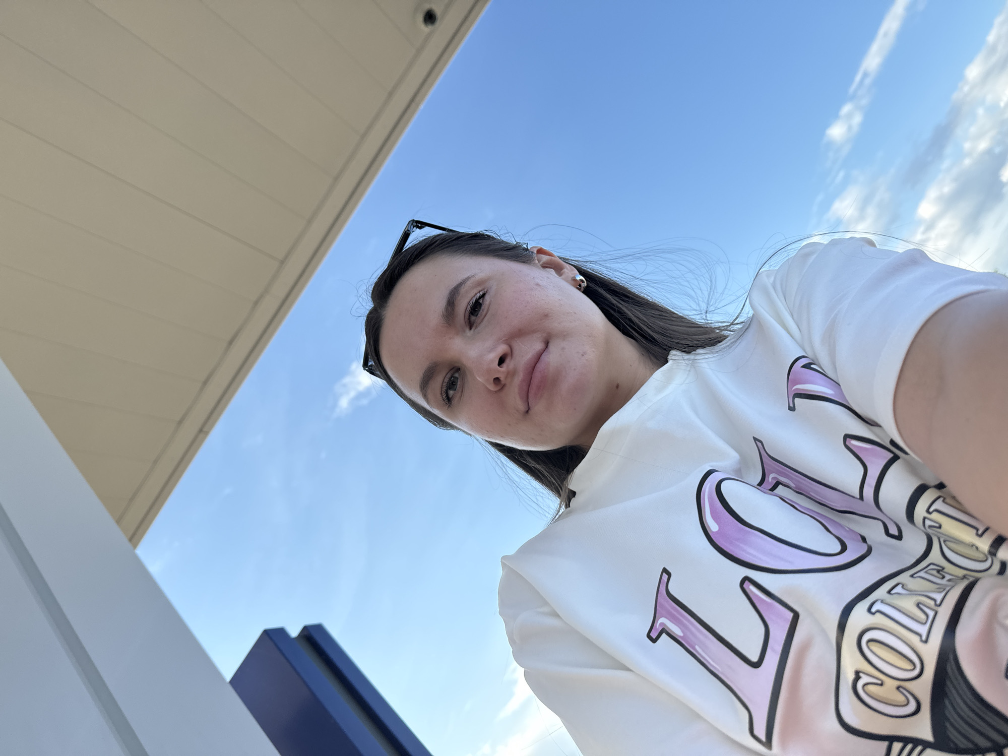 A young woman is taking a selfie outdoors. She has long brown hair, and she's wearing a white t-shirt with the word "COLLEGE" printed on it. Sunglasses are perched on top of her head. The background consists of a clear blue sky with some wispy clouds. A section of a white awning is visible in the upper left corner, as well as a blue and white architectural element in the lower left corner. This scene is located in Bolshiye Vyazyomy, Russia.
