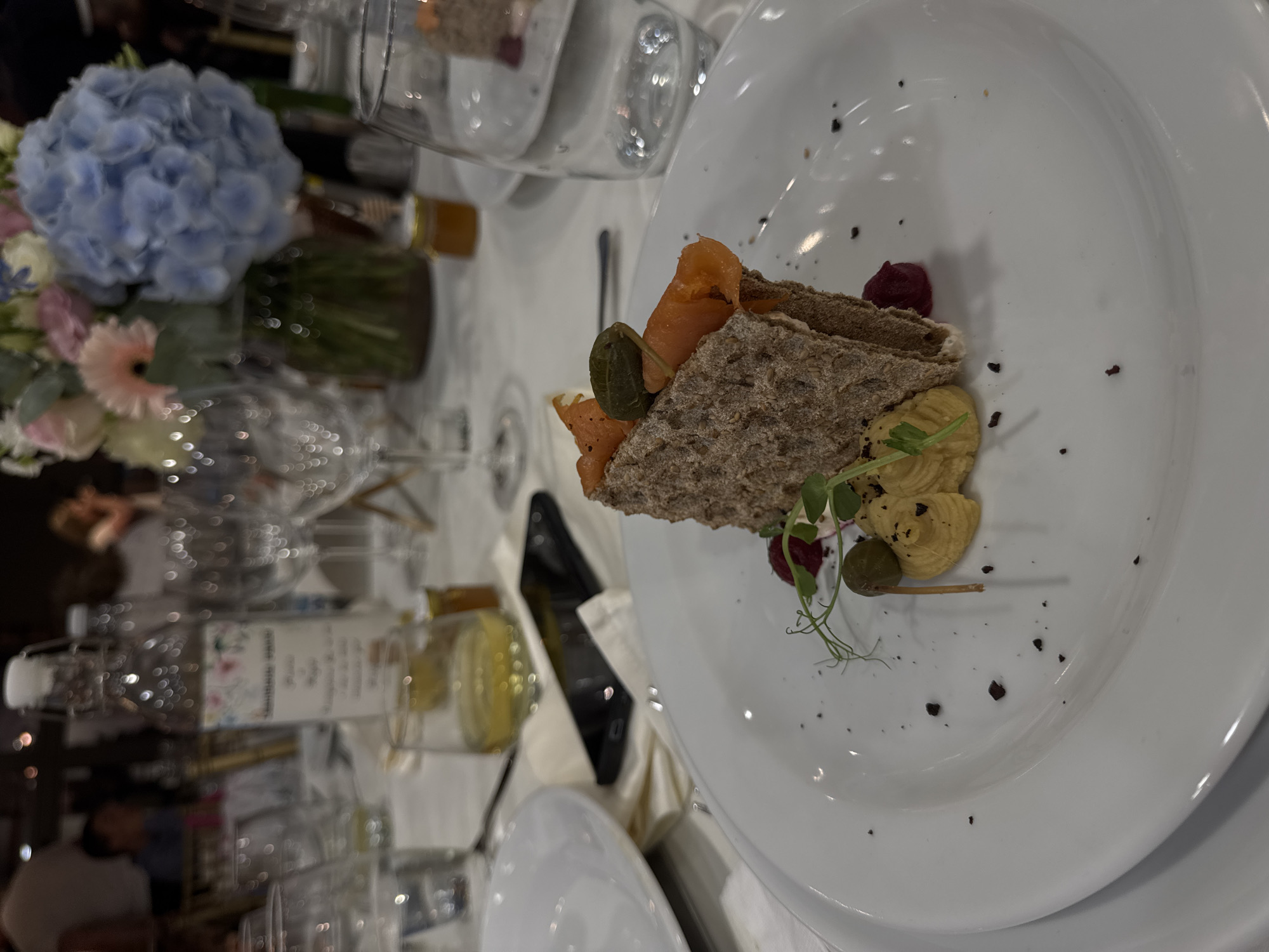 The image depicts a plate of food at a formal event. The main component is a piece of dark bread topped with what appears to be smoked salmon, a green caper, and a red beet garnish. A light-colored, whipped spread is also on the plate, accompanied by microgreens and scattered dark granules.
The plate sits on a white tablecloth alongside a variety of glassware, silverware, and floral arrangements featuring hydrangeas. A black smartphone rests on a napkin near the plate. The setting suggests a wedding or a celebratory dinner, though the location in Mogilovo, Bulgaria is not evident from the visual information provided in the image. No people are visible.