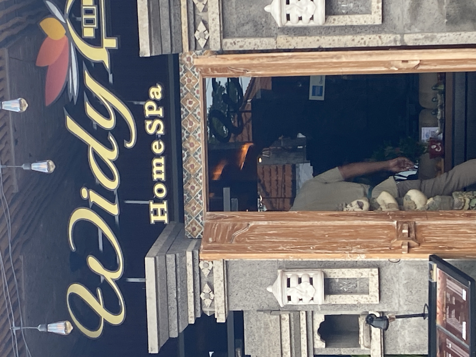 This image displays the exterior of a business named "Didy HomeSpa," likely located in Kuta Selatan, Indonesia. The scene is illuminated by natural daylight.
A prominent dark blue sign features the name "Didy" in elegant gold cursive script, accompanied by a stylized floral or leaf design in red, orange, and white. Below this, "HomeSpa" is written in gold block capitals. Several modern pendant lights with clear glass shades hang from above, illuminating the sign.
The building's facade is constructed from textured gray stone or concrete, adorned with white, intricately carved decorative elements, including stylized lantern-like features and geometric patterns. A wooden-framed window, embellished with patterned mosaic tiles around its border, forms a central part of the facade.
Through the window, reflections of the outdoor environment, such as trees or tropical foliage and potentially a parked scooter or motorcycle, are visible. Inside the spa, a person wearing a light-colored (possibly olive green or beige) top is partially seen, with their arm and torso suggesting they are engaged in an activity within the premises. A row of small, decorative, skull-like objects or sculptures is neatly arranged on a ledge inside the window. Further back, large glass bottles or containers can be dimly discerned. To the lower right, a black screen or monitor on a stand is visible. The architectural style and decorative elements are consistent with the tropical and cultural aesthetics prevalent in Bali.