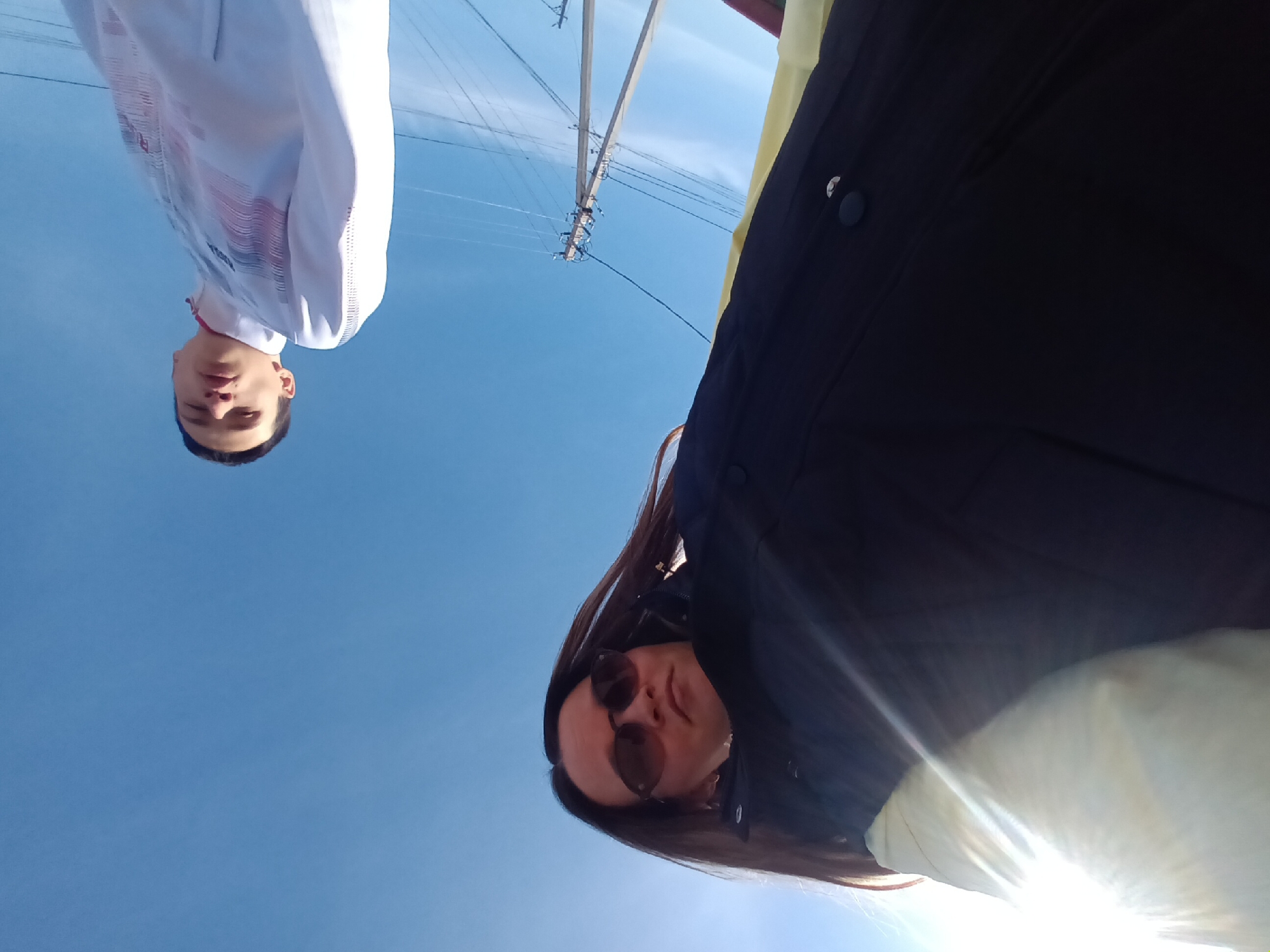 The image shows two people, a woman with sunglasses and a dark jacket, and a man with a white shirt with a red design. The image is upside down and taken from a low angle, looking up at the subjects against a blue sky. A power pole is faintly visible in the background. The location is in the city of Ольховка, Russia.