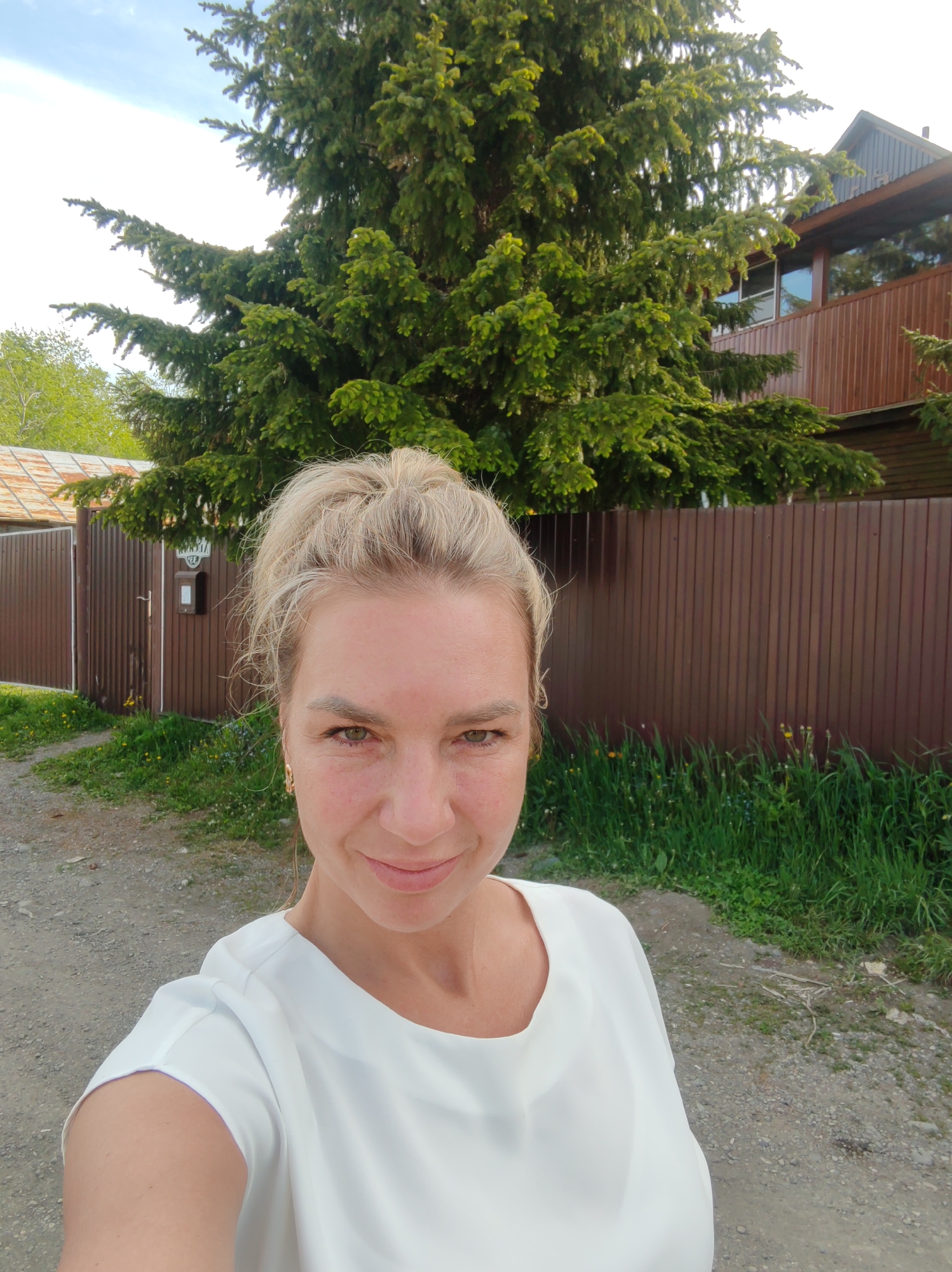 A woman with blonde hair pulled into a bun takes a selfie outdoors. She is wearing a white top. Behind her is a brown fence, a large evergreen tree, and a wooden house. The ground in front of her is a dirt road. There is green grass and some small yellow flowers growing along the fence. The sky is cloudy. The location is the city of Paratunka, Russia.