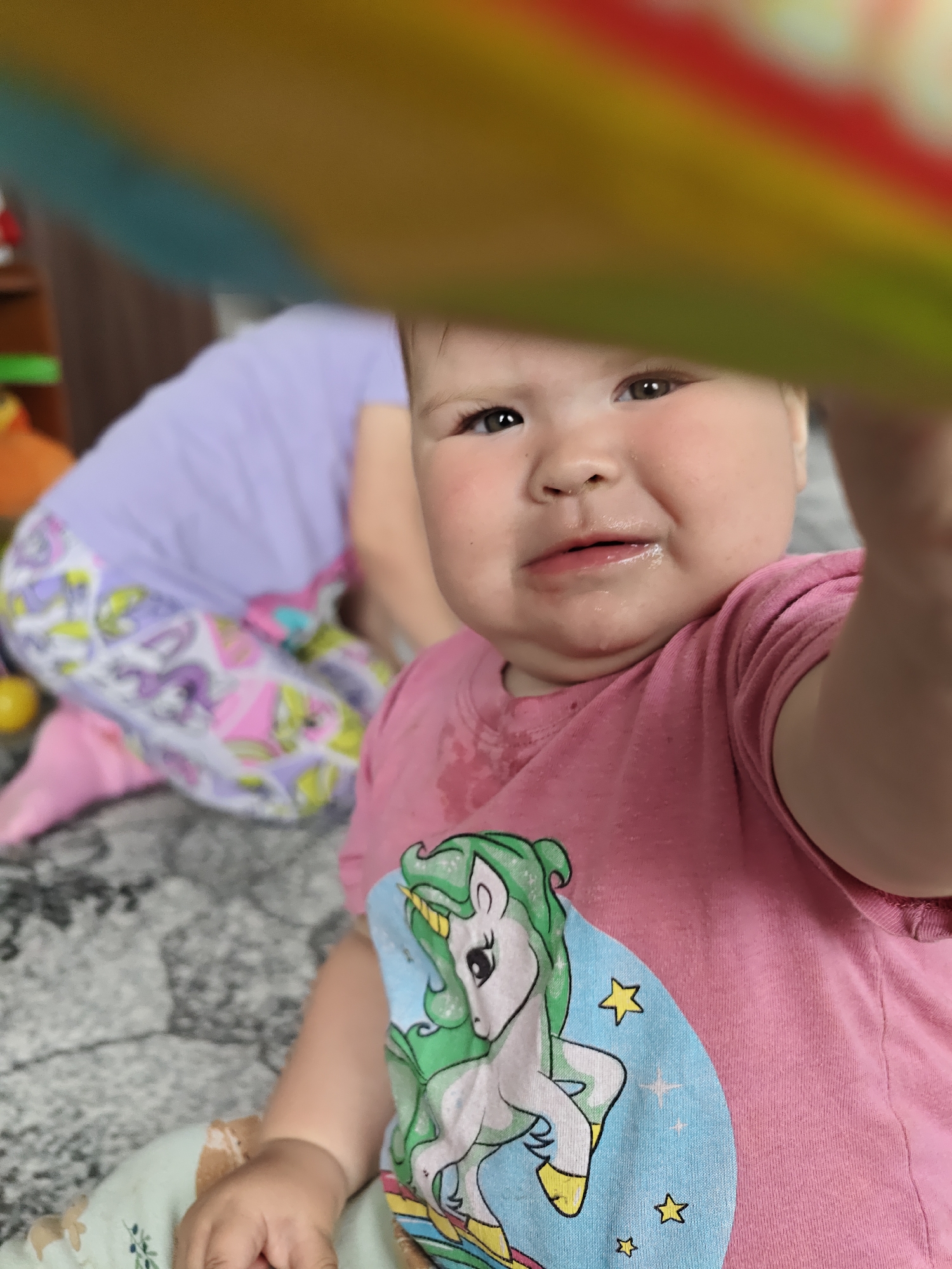 A close-up shot captures a baby girl with tearful eyes and a slightly downturned mouth, suggesting she is crying. She is wearing a pink t-shirt with a unicorn graphic. The shirt appears wet, possibly due to tears or drool. An out-of-focus, rainbow-colored object is in the foreground. Behind the baby, another child wearing a purple shirt and patterned pants sits on a gray carpet. The location appears to be indoors, possibly a home in Уральское, Russia.