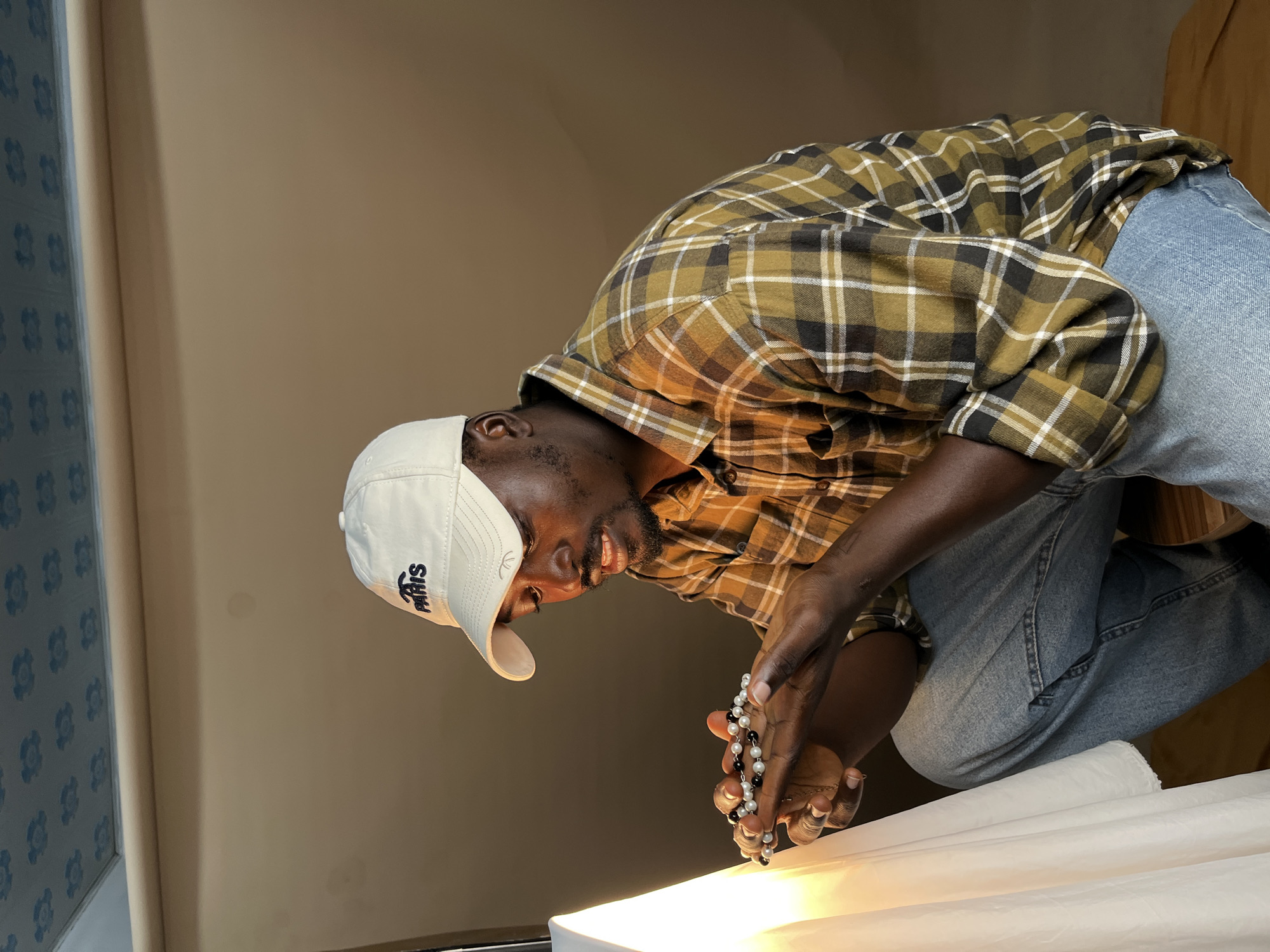 A young Black man wearing a white baseball cap with "PARIS" embroidered on it and a brown, gold, and black plaid shirt is crouching. He is looking down at a necklace or bracelet he is holding in his hands. The necklace appears to be made of black and white beads. He is wearing blue jeans. The background is a plain, neutral-colored wall, with a white sheet of fabric to the left, and a light source creating a bright illumination on the fabric and the man's left side. The ceiling has a blue pattern on it. The scene is indoors. There is no indication of the location being in Gura Top, Nigeria.
