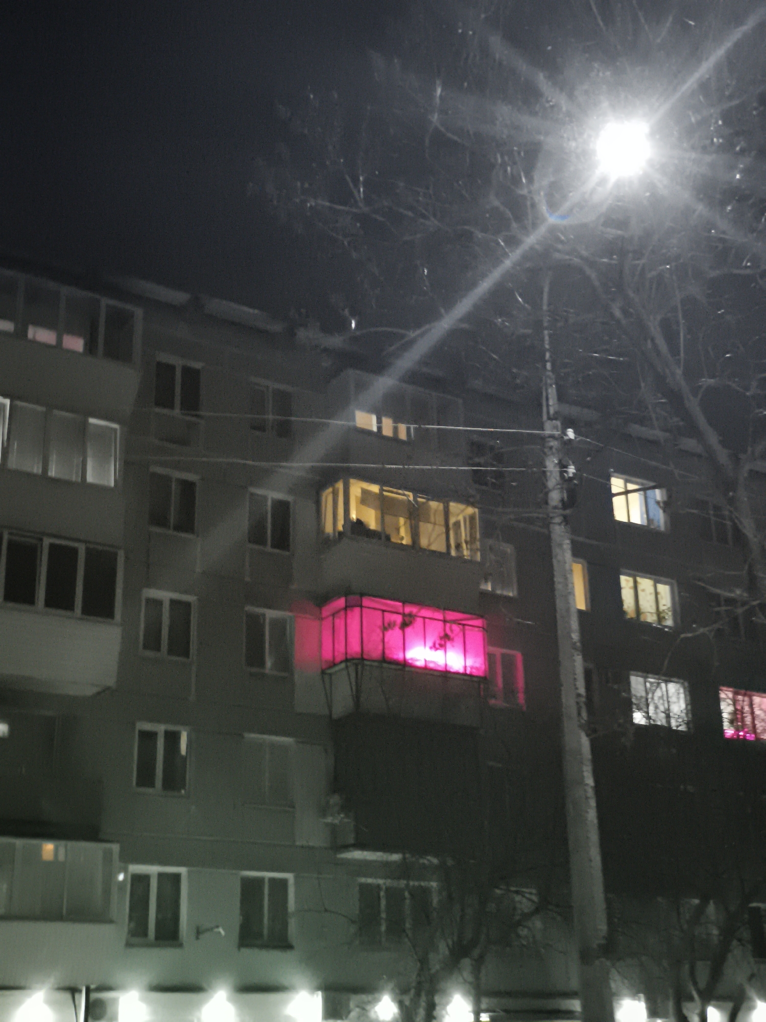 This nighttime photograph, taken in Chaikovsky, Russia, displays a section of a multi-story residential building. The building's facade features numerous windows, some dark, others emitting light from within. A prominent enclosed balcony on an upper-middle floor is brightly illuminated with a distinct magenta-pink light. Directly above this, another enclosed balcony emits a warm yellow-orange glow. Various other windows across different floors show ambient interior lighting. To the right, a utility pole stands vertically, topped with an intensely bright streetlamp that produces a significant starburst effect. Bare tree branches, indicative of a dormant season, are visible in the upper right quadrant and partially obscure parts of the building. No individuals or specific actions are discernible within the frame.