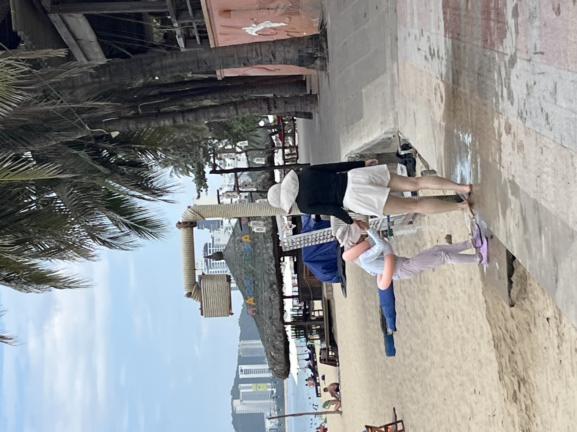 This image captures a scene on a beach in Nha Trang, Vietnam, with a view of the city skyline in the background. In the foreground, a person wearing a white wide-brimmed hat and a black long-sleeved shirt with white shorts stands on a wet paved area facing away from the camera. To their left, another person, dressed in a light blue shirt and light purple pants, is interacting with an inflatable pink ring, which is positioned around their torso and extends horizontally. The person in the inflatable ring appears to be bent over. The beach is sandy, with palm trees lining the area. A thatched-roof structure, possibly a bar or restaurant, with the words "HAPPY BIRTHDAY" visible on its awning, is situated further down the beach. Other beachgoers and umbrellas are visible in the distance. The overall atmosphere suggests a casual day at the beach.