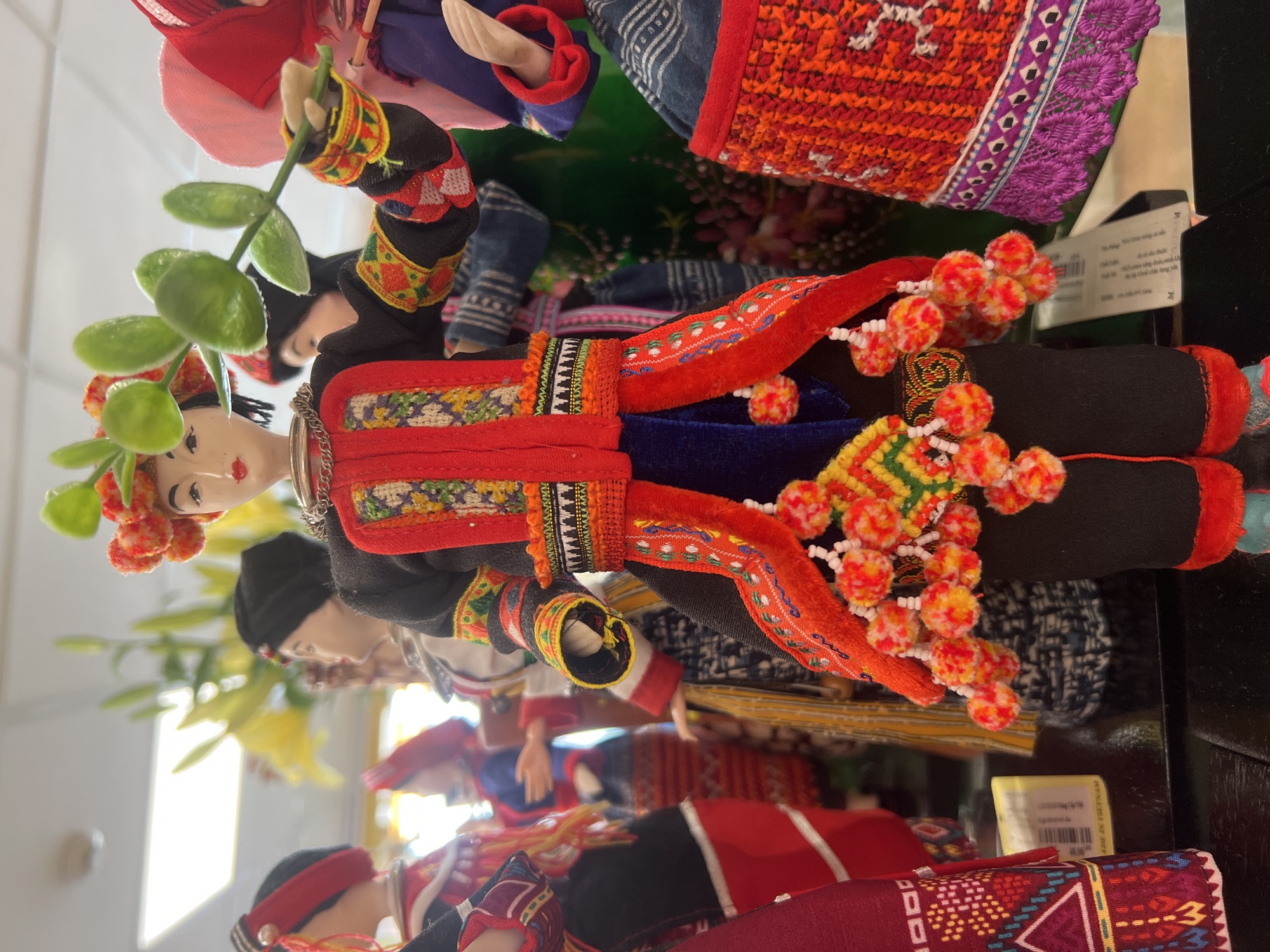 The image shows a collection of traditional Hmong dolls displayed for sale, likely in a market or souvenir shop in Hà Nội, Vietnam. The dolls are intricately dressed in colorful, embroidered clothing featuring geometric patterns and decorative tassels. One doll in the foreground is holding a green leafy branch, while others stand behind, showcasing their vibrant attire. The background is slightly blurred, revealing more dolls and hints of yellow flowers, suggesting a retail setting. The lighting appears bright, possibly from overhead fixtures.
