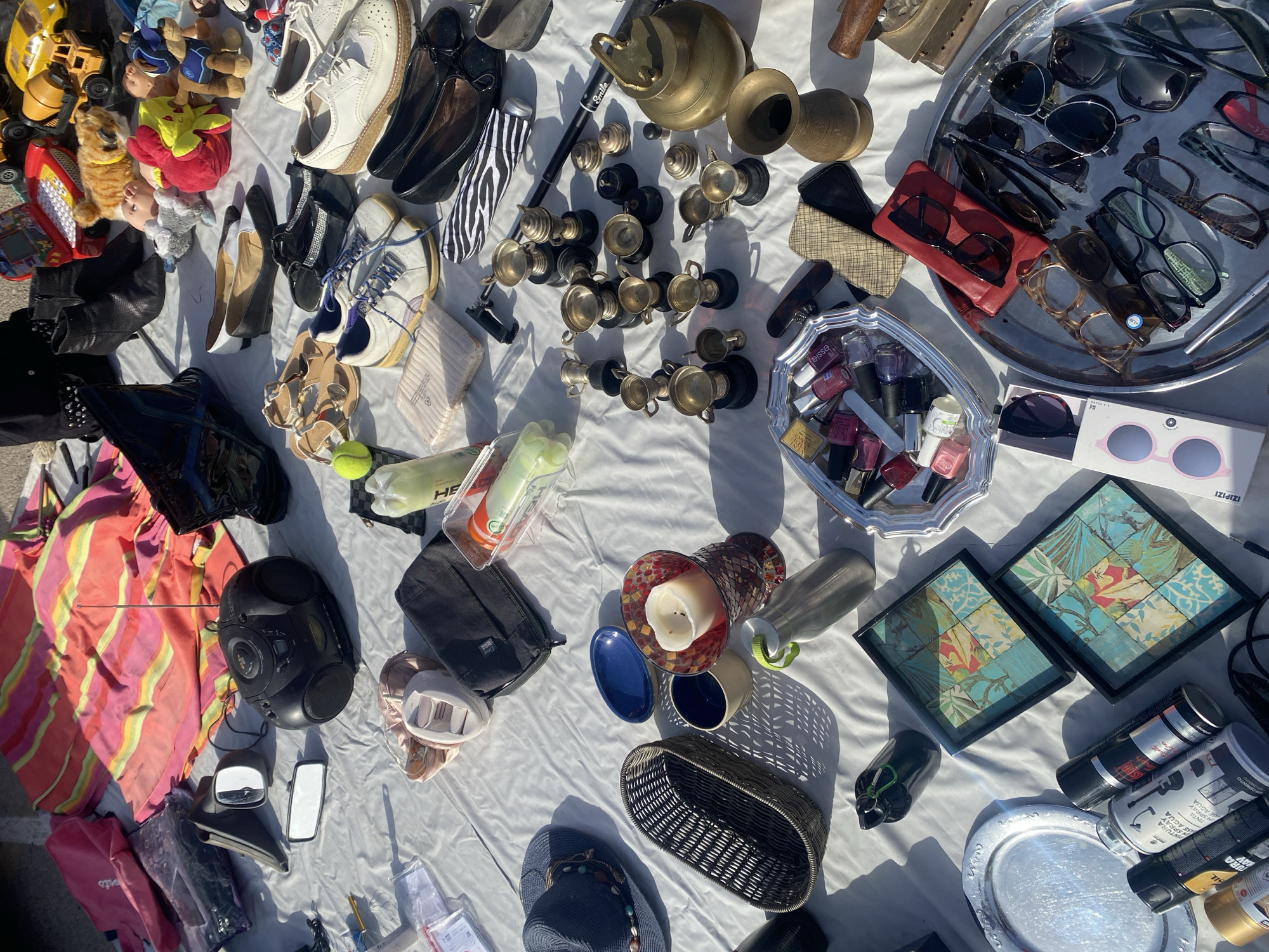 The image depicts a flea market stall set up outdoors on a white tarp. The stall displays a variety of items including clothing, shoes, accessories, decorative items, and a boombox. In the background, a brightly colored blanket with stripes in shades of pink, yellow, and orange is visible. The ground appears to be paved, suggesting an urban or semi-urban outdoor setting. The lighting indicates it is daytime, with shadows cast by the objects.
Some of the discernible items include:
* A black boombox with an antenna.
* A pair of car mirrors.
* Various shoes, including white sneakers, black flats, and sandals.
* A blue hat with beaded trim.
* A woven basket.
* A candle in a decorative holder.
* Several small trophy cups.
* A brass teapot or pot.
* A tray filled with nail polish bottles.
* A selection of eyeglasses and sunglasses.
* A metal tray.
* Several spray cans, some labeled with Spanish text.
* Two framed pictures with a decorative pattern.
The overall impression is of a typical outdoor market scene where pre-owned goods are being sold. The presence of Spanish labeling on some items, combined with the prompt's mention of Manilva, Spain, suggests this market is located in that region.