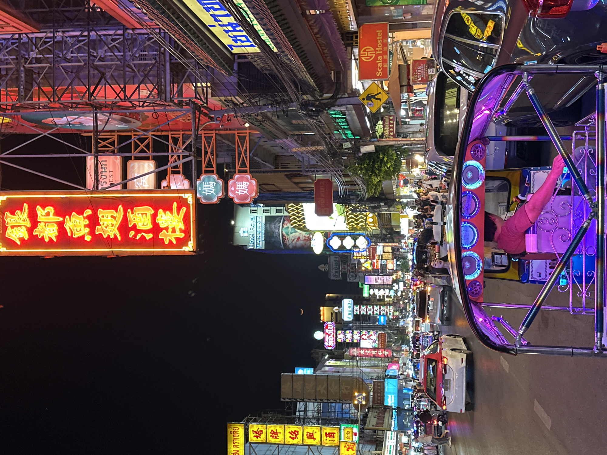 This image captures a bustling street scene at night in Bangkok, Thailand, characterized by a profusion of bright neon signs in Thai and Chinese, towering above the street. A tuk-tuk, adorned with neon blue lights, sits parked on the right side of the frame. A driver is visible in the vehicle. The street is busy with pedestrians and vehicles. The signs advertise restaurants, hotels, and other establishments. A partial moon can be seen high in the upper central part of the image. A Scala Hostel sign is visible above and to the right of the tuk-tuk. Overhead, a tangle of electrical wires adds to the urban environment.