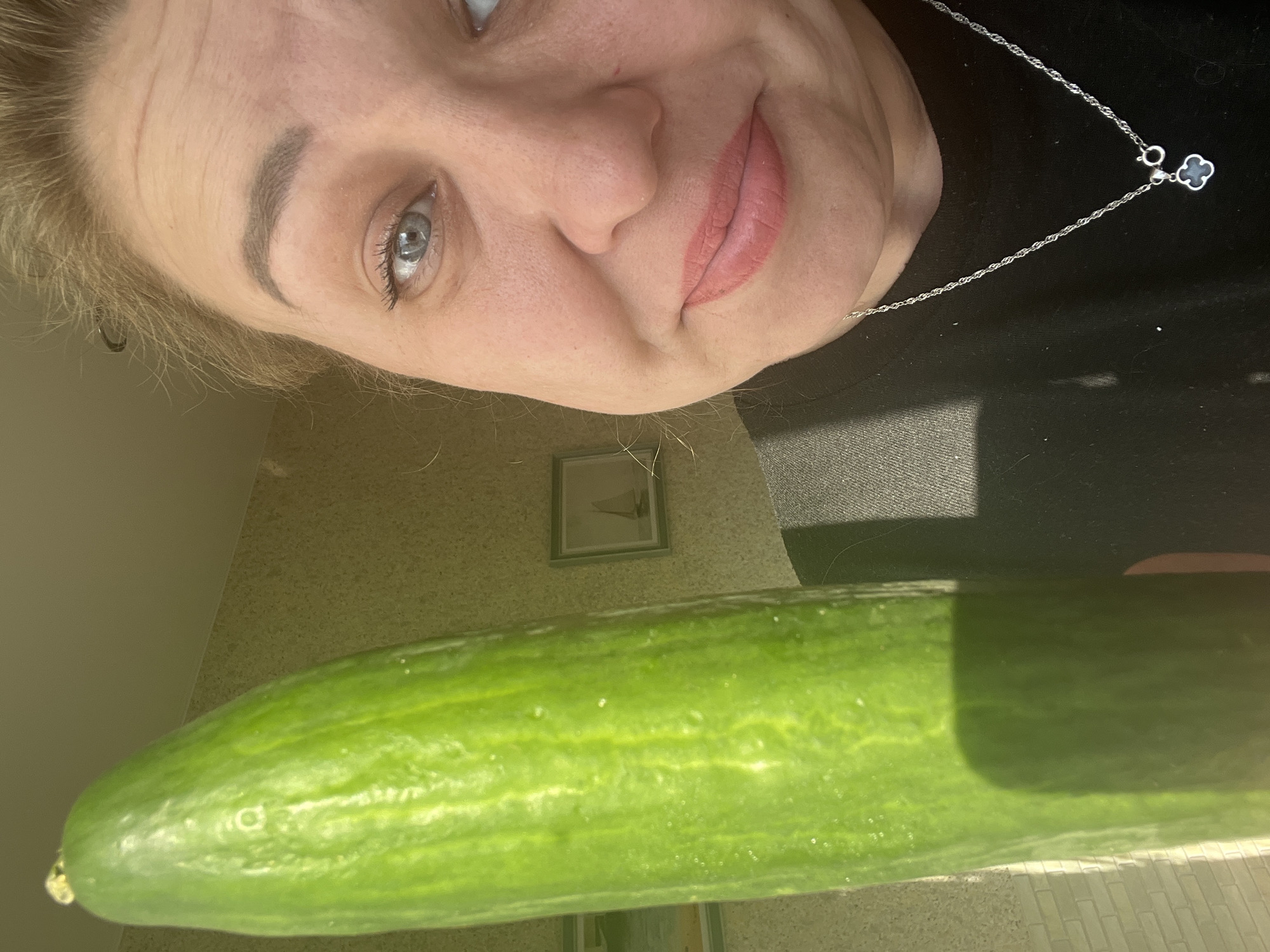 A woman is holding a large green cucumber in front of her face. The cucumber appears to be freshly washed and is the focal point in the foreground. The woman has fair skin, light-colored hair, and blue eyes. She is wearing a black top and a silver necklace with a clover-shaped pendant. The background includes a light-colored wall and a framed picture. There are tiles at the bottom of the frame. The location in the city of Chaikovsky, Russia, cannot be determined from the visual data provided.