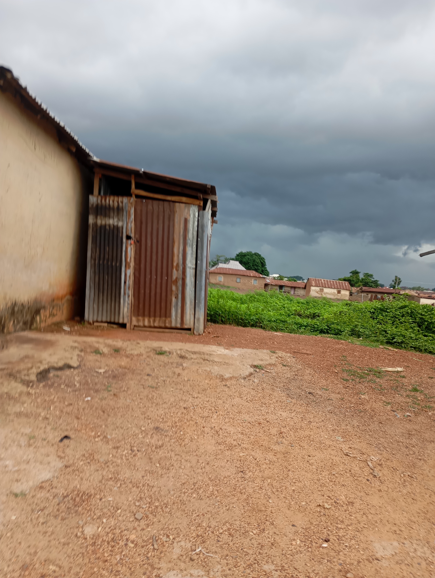 The image depicts a scene in Shendam, Nigeria, under a heavily clouded sky. In the foreground, a dirt path leads towards a building with a rough-textured, light-colored wall and a corrugated metal door. The door appears to be ajar, revealing a dark interior. To the right of the building, a patch of lush green vegetation is visible, bordering a collection of houses with rust-colored roofs. The houses are modest in construction, suggesting a rural or semi-urban setting. The overall impression is one of an overcast day, possibly before or after a rain shower, in a developing area of Shendam. There are no people visible in the image.