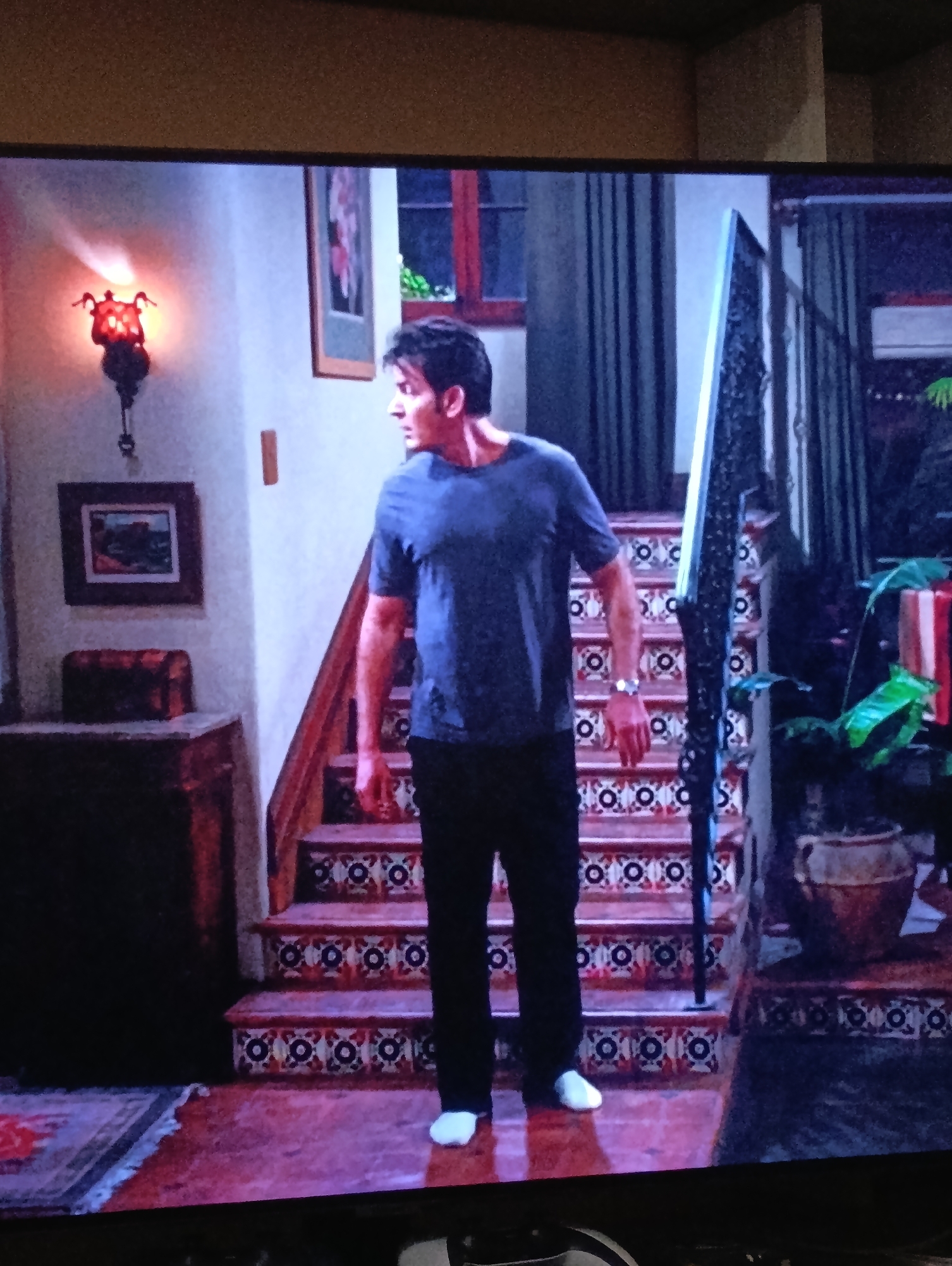 A man with short dark hair, wearing a blue t-shirt, black pants, and white socks, stands on the first step of a staircase. He is looking to his left. The staircase has decorative, patterned tiles. A black railing is on the right side of the staircase. A plant in a pot sits at the base of the stairs. To the left of the stairs is a dark wooden cabinet with a red decorative chest on top. A framed picture hangs above the cabinet. A wall sconce with a red light is above the framed picture. A window is visible behind the man, and a curtain hangs beside it. The scene appears to be a still from a television show or movie. The location is not in the city of Plovdiv, Bulgaria.