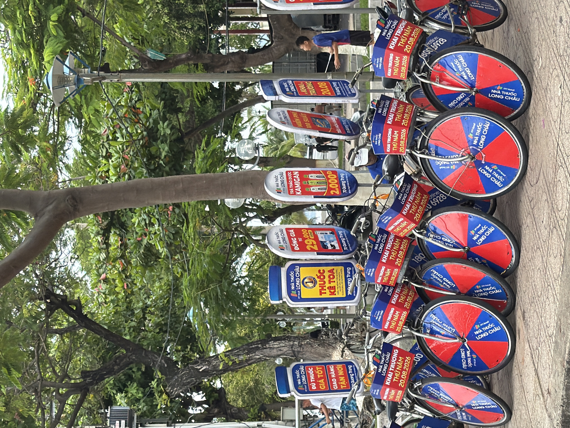 The image depicts a street scene in Nha Trang, Vietnam, with several bicycles parked in the foreground. The bicycles have blue and red patterned wheels and blue baskets with signage. Prominent on the bicycles are signs in Vietnamese, one shaped like a large pill bottle advertising "LONG CHAU" and another with the text "KHAI TRƯỜNG THỨ NĂM 20.08.2026". In the background, trees, a lamppost, and several people are visible. One man in a blue shirt is holding a stick near a bicycle, while other individuals are walking in the distance. The setting appears to be an outdoor public space, possibly a sidewalk or park area.