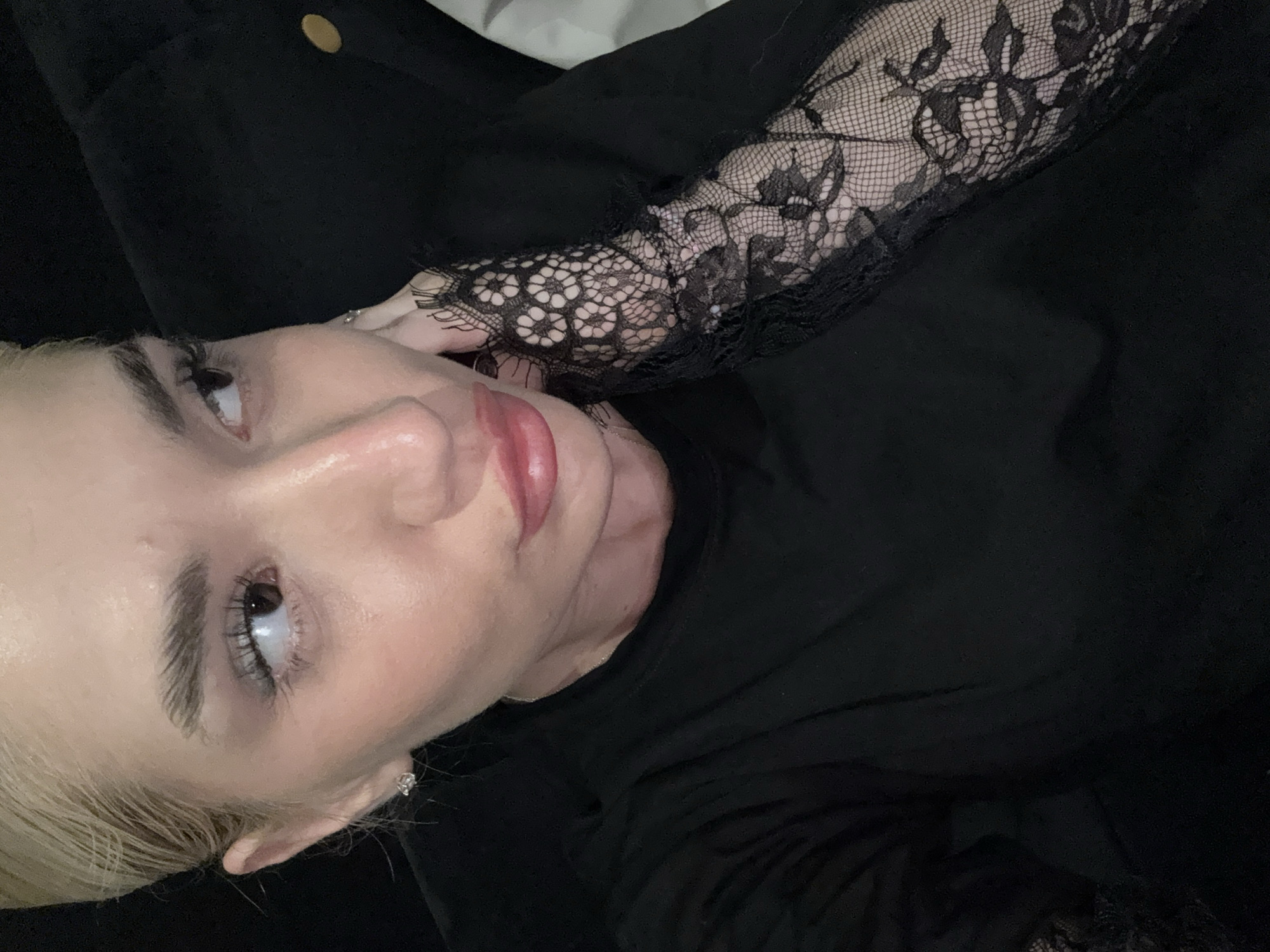 A close-up, high-angle portrait captures a person, likely a woman, with fair skin and light-colored hair, possibly blonde. The subject is seen from a slightly tilted, vertical perspective, looking upwards and to their right. Their left hand gently touches their face near the jawline.
The person has distinct eyebrows, mascara-enhanced eyelashes, and lips colored with a soft red or pink lipstick. A small, sparkling earring is visible in their left earlobe. They are dressed in a plain black long-sleeved top or dress. Their right arm (on the left side of the image) is covered by a black lace sleeve or glove, featuring an intricate floral or abstract pattern, which adds a decorative element to the dark attire.
The background is predominantly dark, suggesting an indoor setting, possibly a dimly lit room or against dark upholstery, with a hint of a golden button or stud visible in the upper left corner. The lighting appears soft, highlighting the contours of the subject's face. The expression is neutral and somewhat contemplative. There is no visible text in the image.
The overall aesthetic conveys a sense of elegance or subtle drama, emphasized by the dark clothing and delicate lace. The location context for this media file is Sofia, Bulgaria.