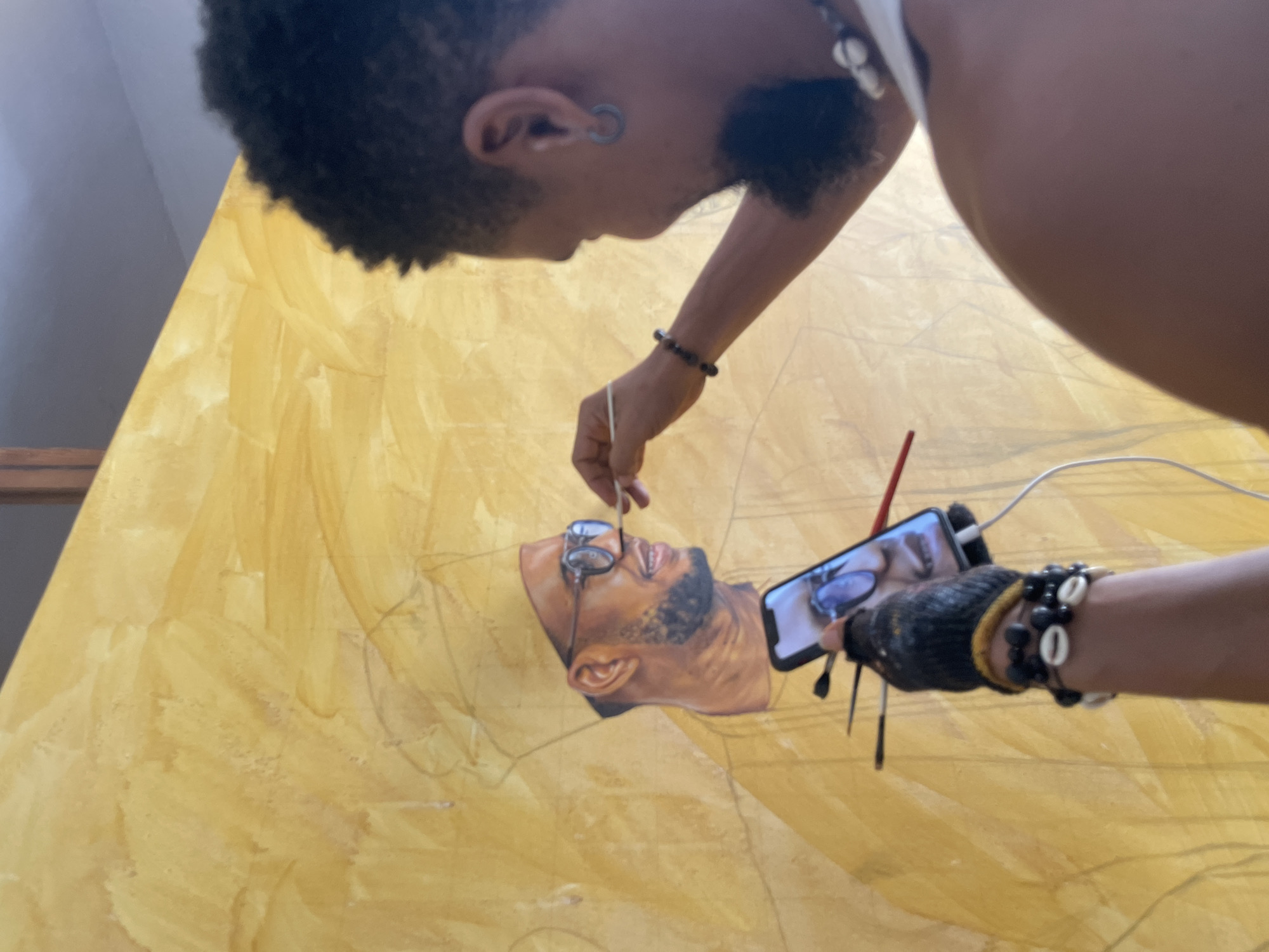 A person with dark skin, short curly hair, and earrings is painting a portrait of a man with a beard and glasses on a large yellow canvas. The person holds a thin paintbrush in their right hand, applying paint to the glasses of the portrait. In their left hand, they hold a smartphone displaying a close-up of the same area of the painting, presumably as a reference. The person wears bracelets on both wrists. The canvas appears to be resting on an easel. The location is indicated as Ado, Nigeria, although no specific landmarks or features confirming the city are visible.