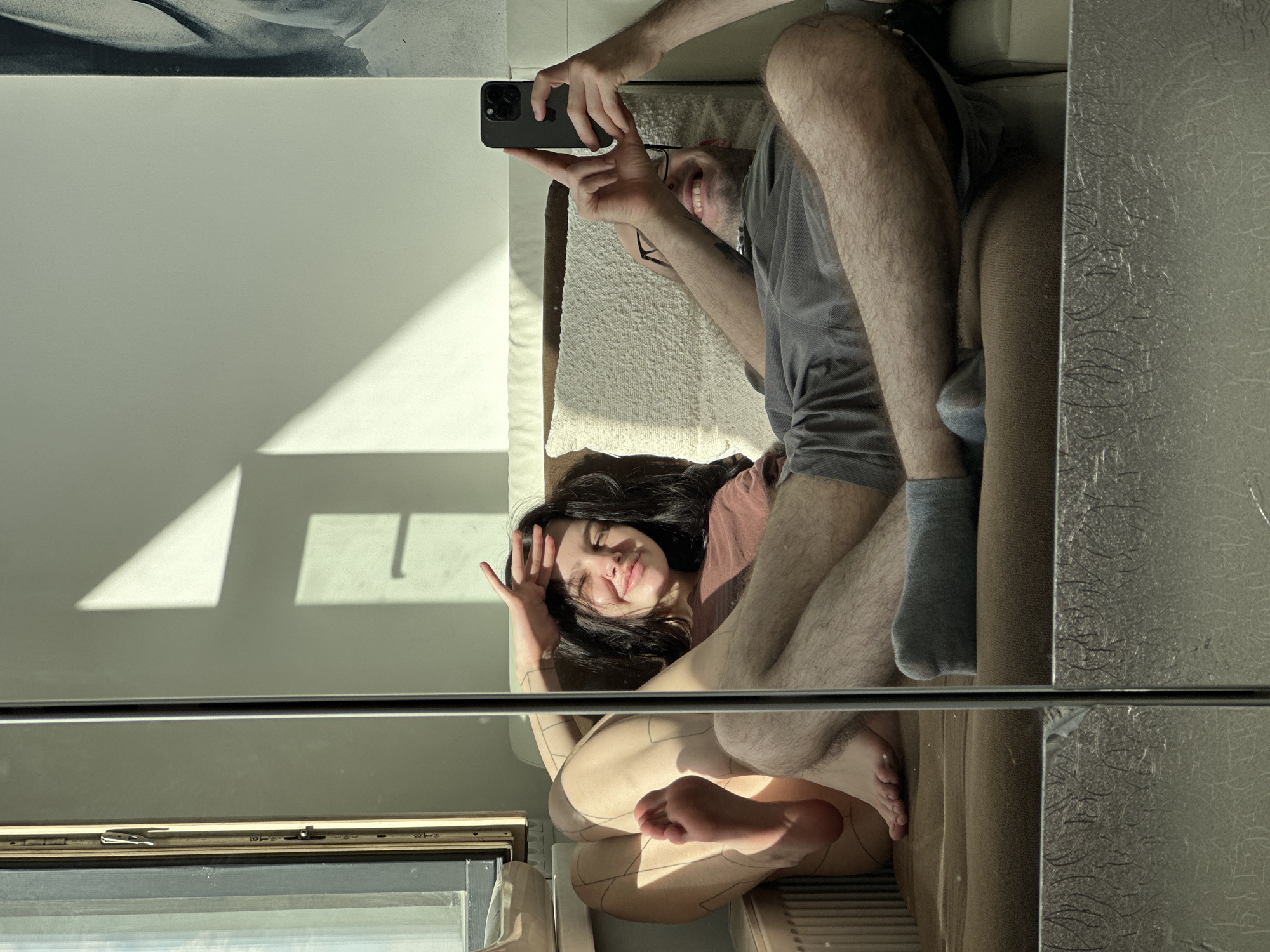 The image shows a reflection in a mirrored wall. A woman with dark hair is lying on a couch, posing for a picture taken by a man with glasses holding a smartphone. The reflection includes a window with sunlight and a radiator. The location is not discernible from the image.