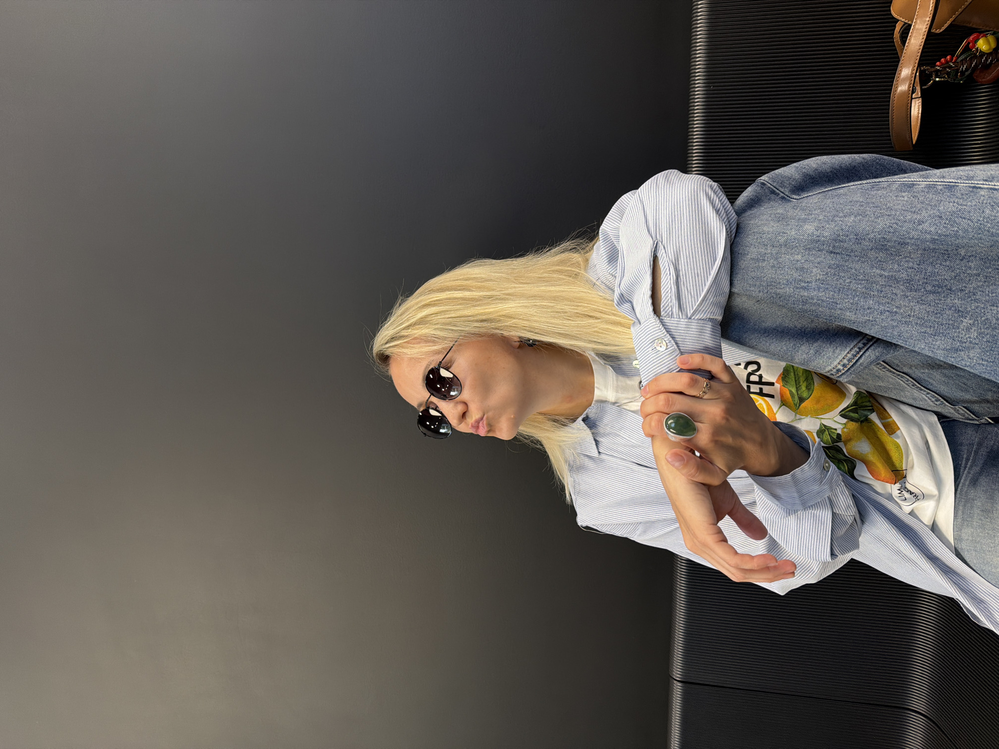 A blonde woman with sunglasses is sitting on a black bench. She is wearing a blue and white striped shirt over a white t-shirt with a lemon print. She has a large green ring on her finger. The background is a dark gray wall. The image was taken in Izhevsk, Russia.