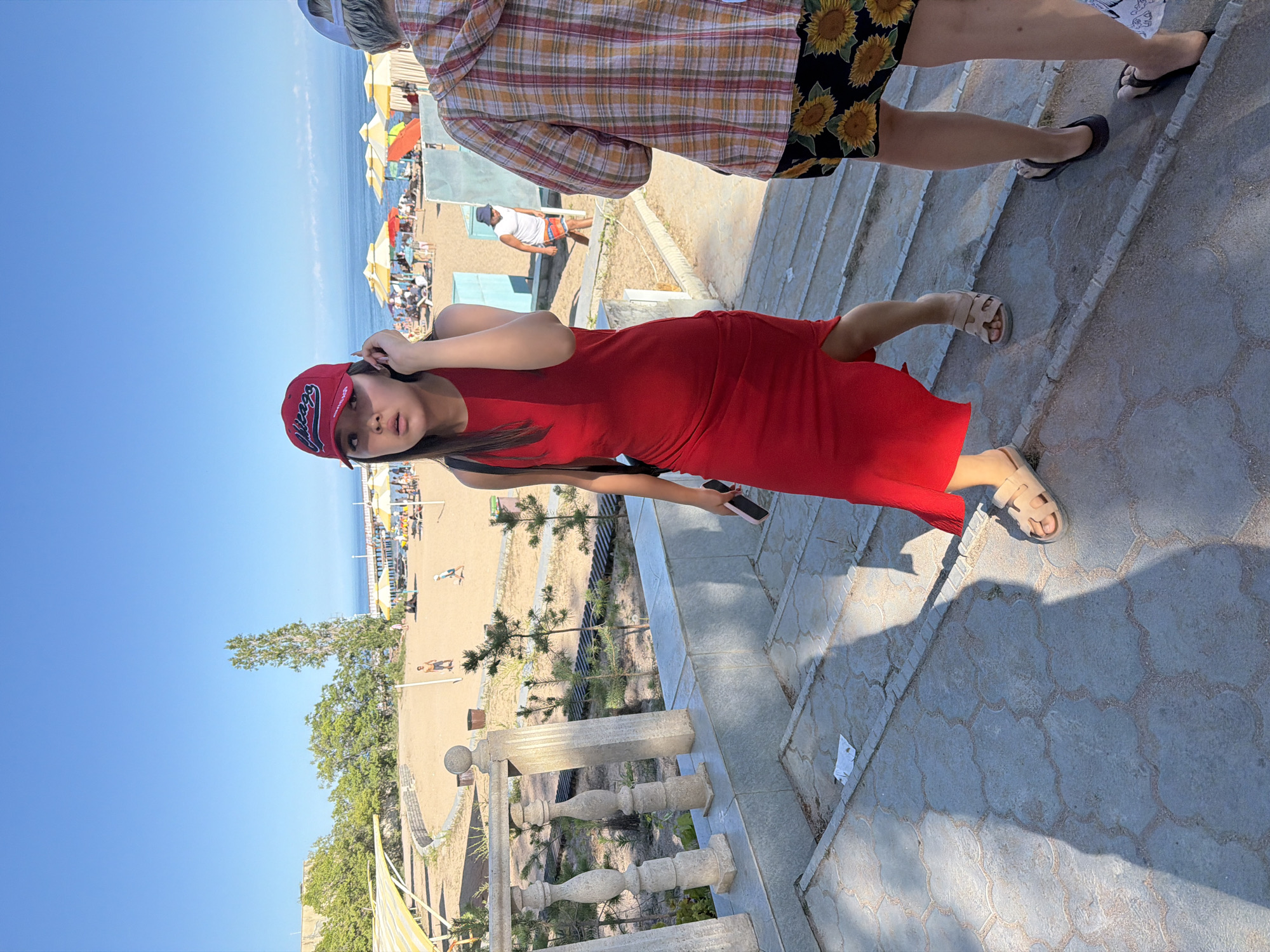 The image shows a sunny day at a beach with a clear blue sky. In the foreground, a young woman in a red dress and a red baseball cap walks down a stone staircase. She is holding a smartphone in her right hand and her left hand is on her ear. To her right, another person is walking up the stairs, wearing a plaid shirt and a skirt with a sunflower pattern. In the background, a beach with sand and umbrellas is visible. People are seen relaxing on the beach and swimming in the ocean. The scene appears to be in a resort or vacation area.